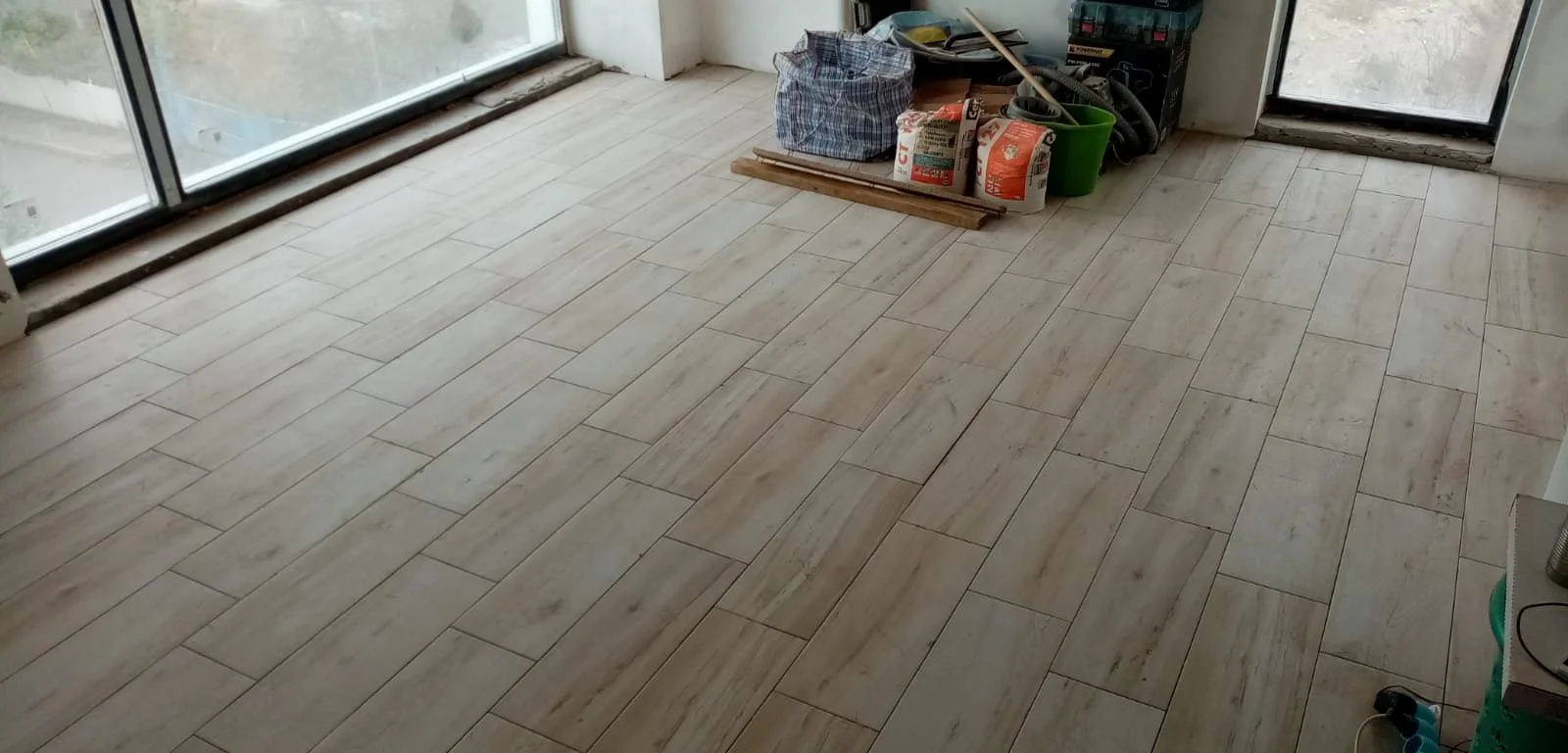 Unfinished room with light-colored wooden floor tiles, large glass sliding doors on the left, and construction tools and materials near the back wall.
