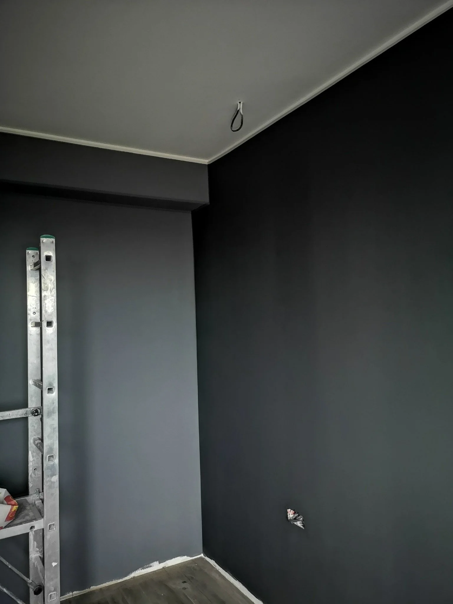 Interior room with dark gray and black walls, ladder, and a hanging pull string on the ceiling, with a small torn piece of wallpaper on one wall.