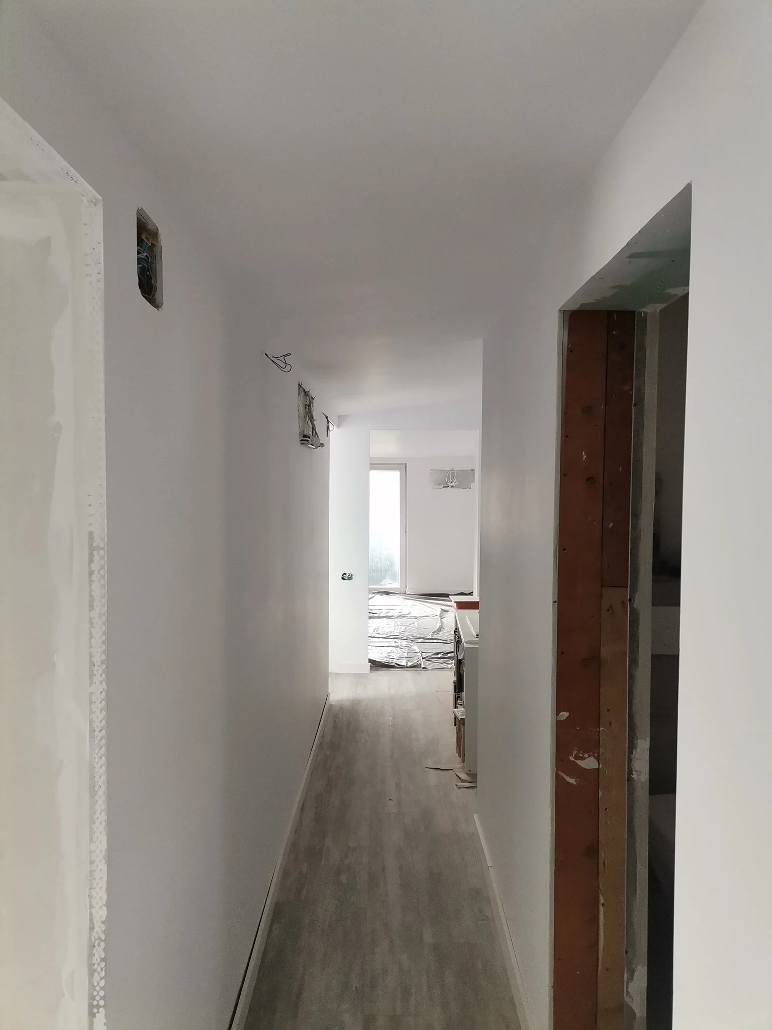 Long narrow hallway under construction with unpainted walls, wires hanging from electrical boxes on the ceiling, and a plastic-covered floor, leading to a window at the end.