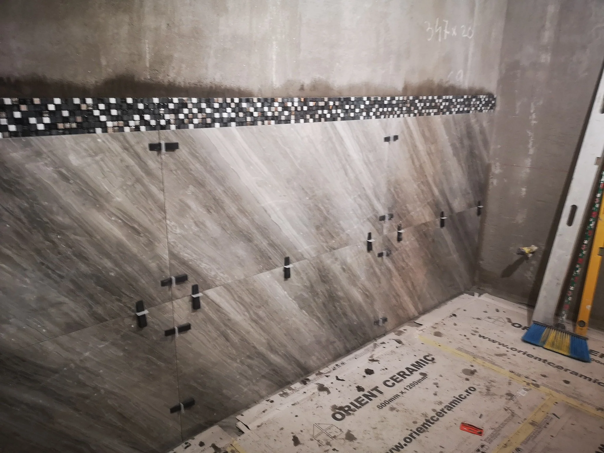 Bathroom wall with partially installed tiles, black and white mosaic tiles in a strip along the wall, construction tools and materials on the floor, including a level, visible at a construction site.