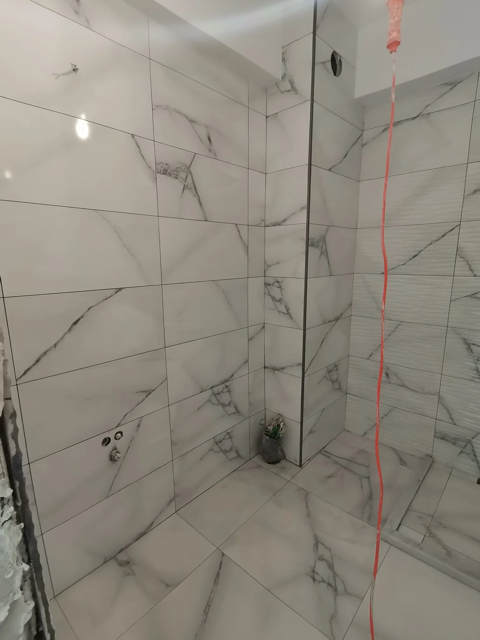 A partially finished bathroom shower with marble tile walls. There is a wire hanging from the ceiling, and an open space where a showerhead or fixture may be installed.