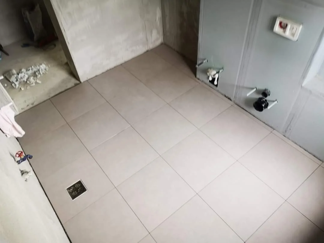 An unfinished room with beige tiled floor, exposed plumbing, and electrical outlets, indicating ongoing construction or renovation.