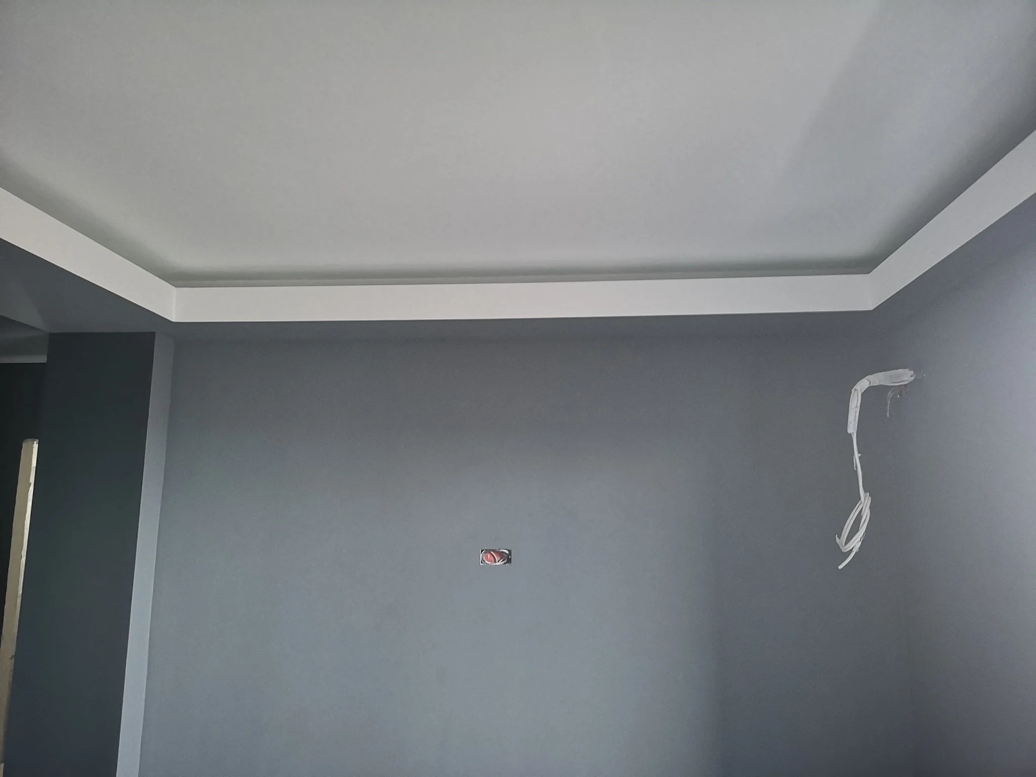 Interior ceiling of a room with white trim, a gray wall, an electrical outlet, and exposed wiring hanging from the ceiling.