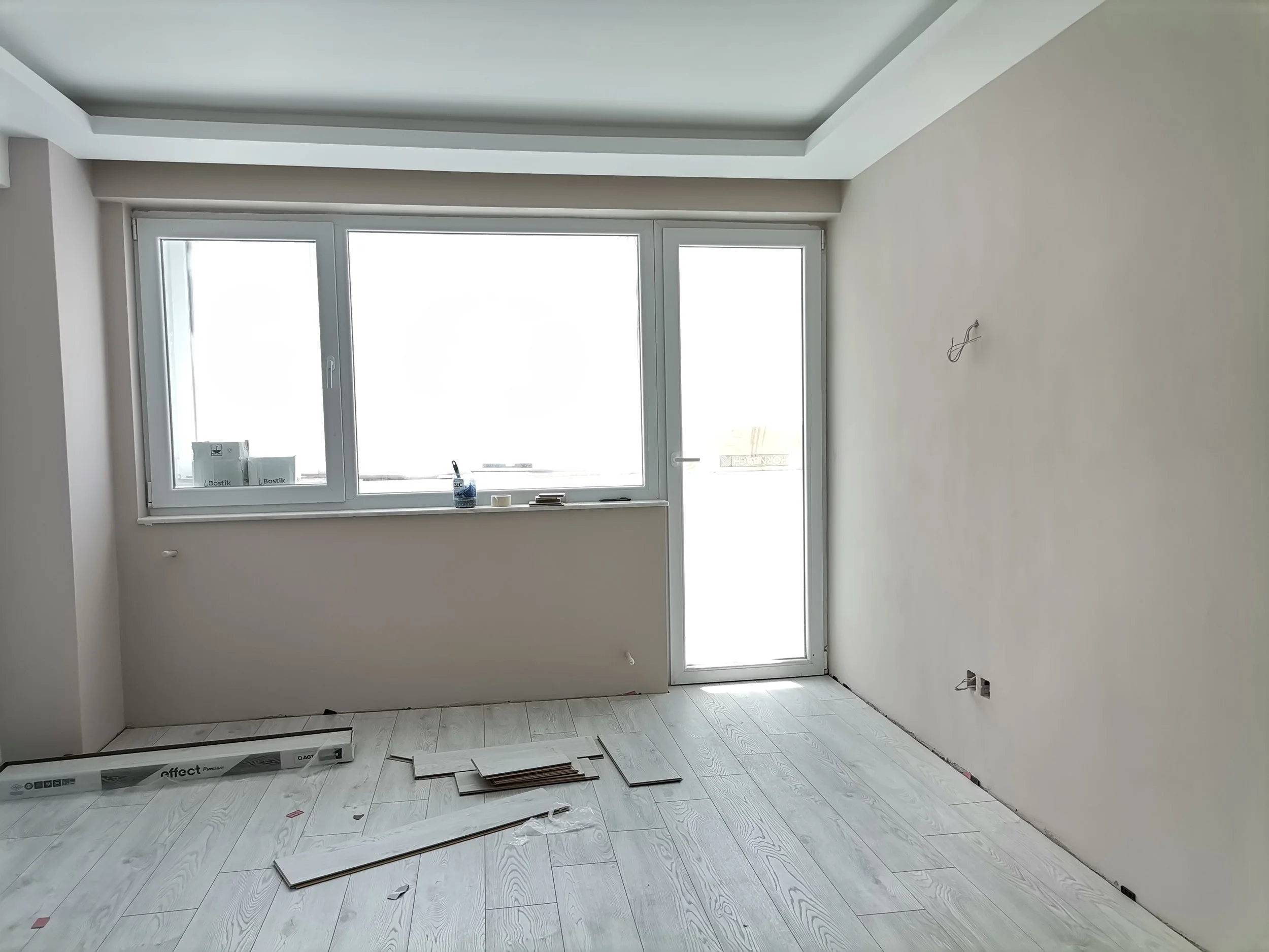 An empty room under renovation with white walls and a large window. Tools and flooring materials are on the floor, suggesting ongoing work.