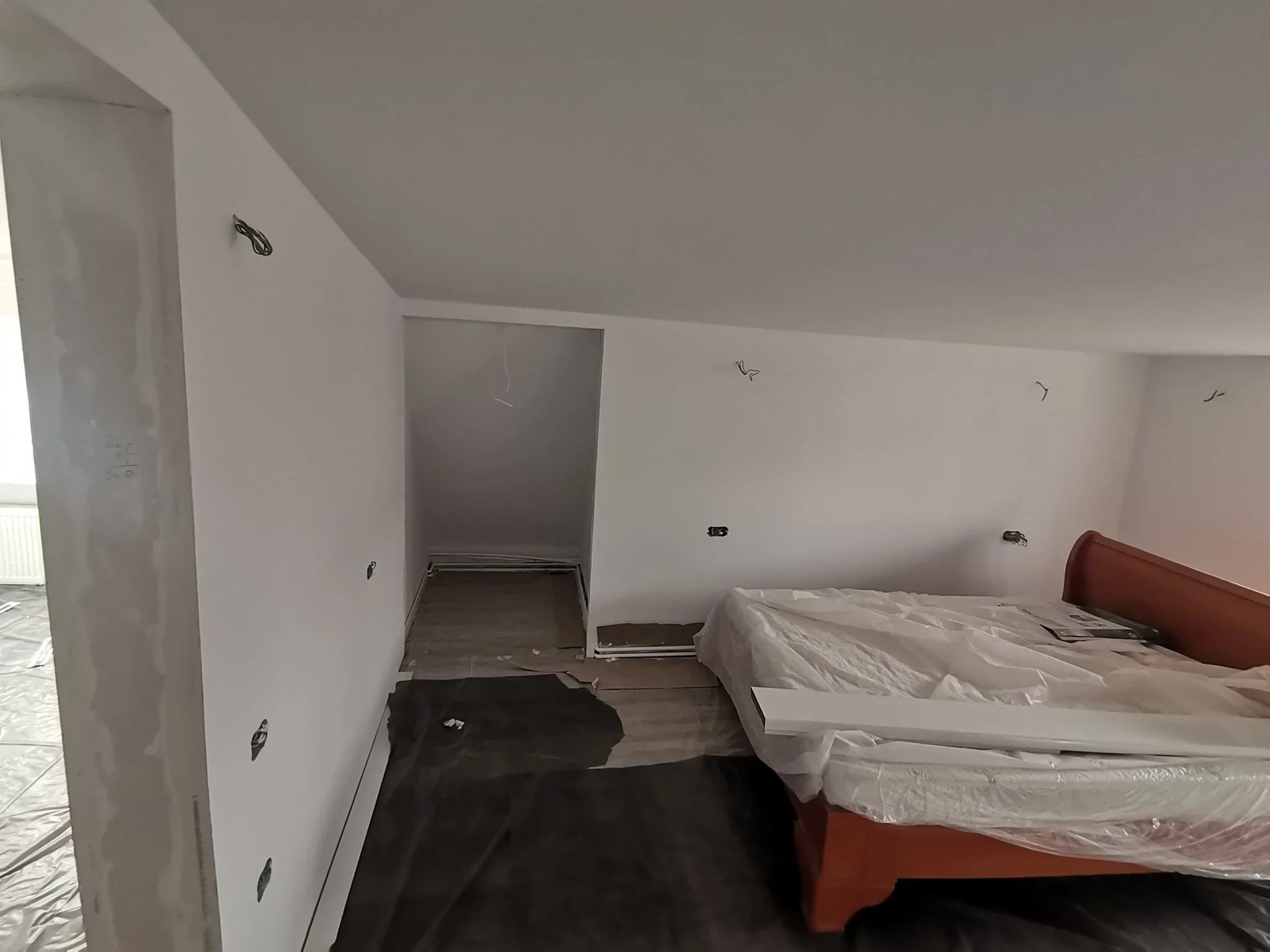 A room under construction or renovation with white walls, some black wires hanging from the ceiling, a bed with a white plastic cover, and scattered construction debris on the dark wood floor.