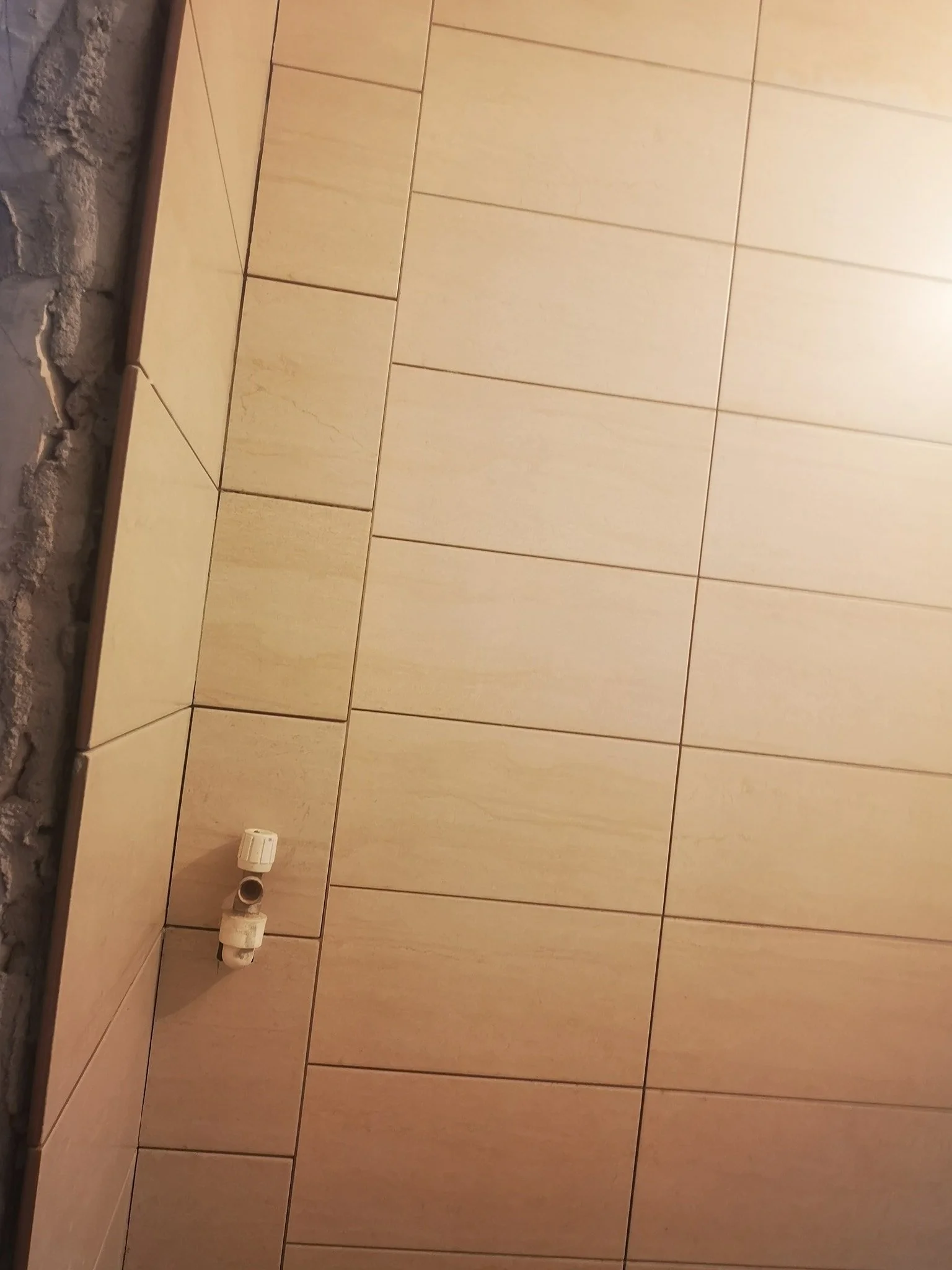 Unfinished bathroom wall with beige tiles, exposed cement and plumbing fittings.