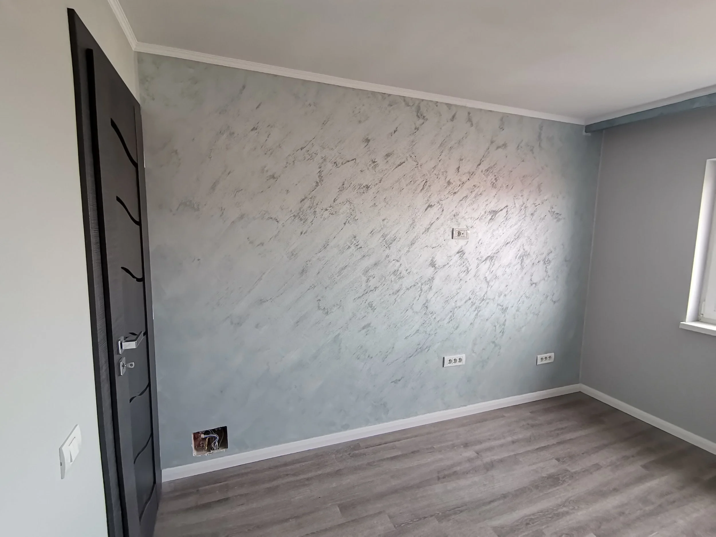Empty room with gray textured wall, gray ceiling, and dark wood flooring. Black door with decorative wavy lines, and multiple electrical outlets on the wall.