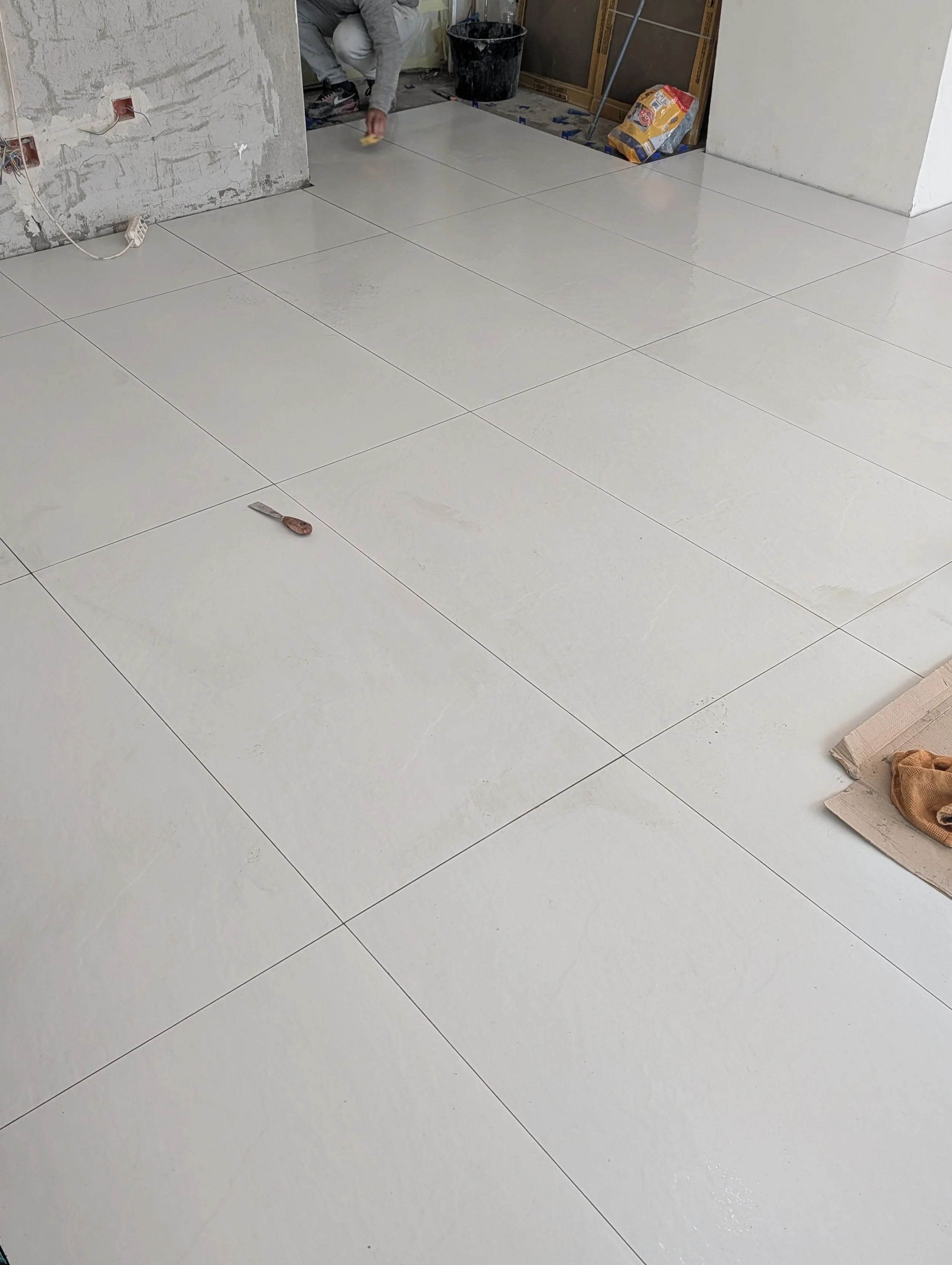 Person installing large white ceramic tiles on a floor in a room under construction or renovation.