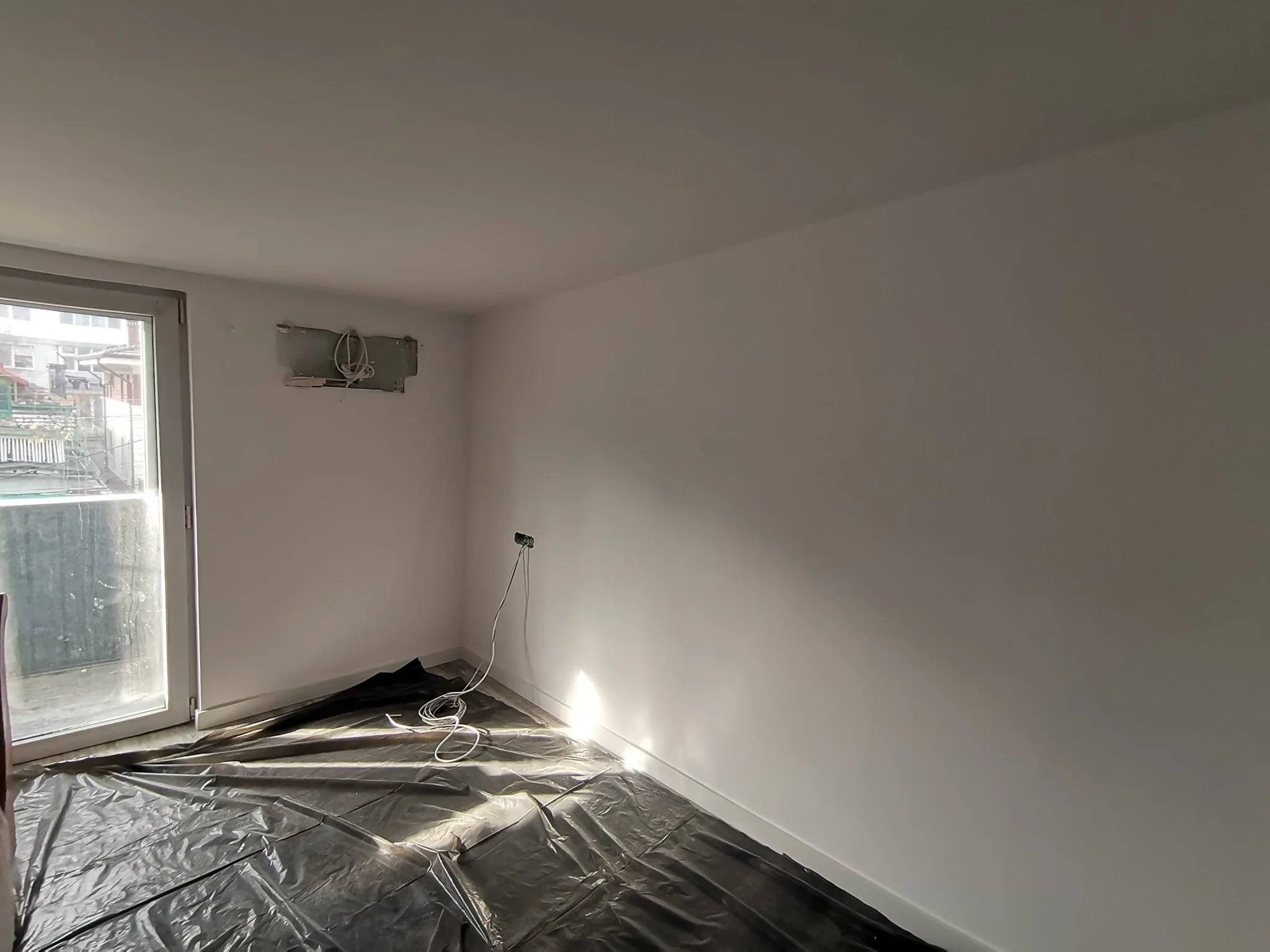 Empty room with white walls, a sliding glass door, and a black plastic sheet on the floor. There are wires hanging near an unfinished wall outlet.
