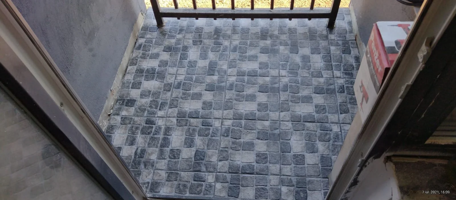 Small outdoor balcony with gray brick patterned tile flooring, black metal railing, and a cardboard box on the right side.