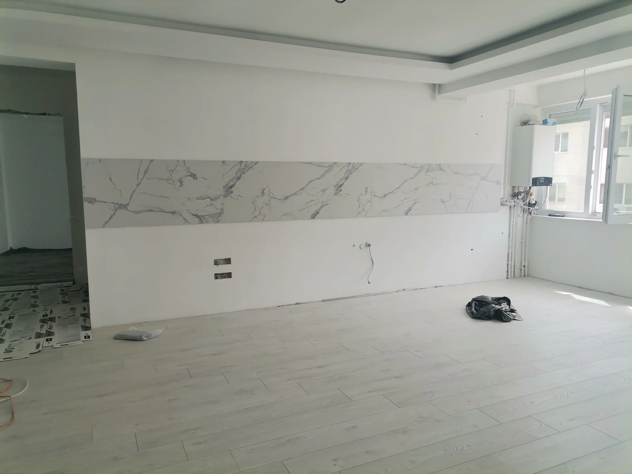 Empty room with white walls, light-colored wooden flooring, and large windows. There are construction materials and plastic bags on the floor, indicating ongoing renovation or construction work.