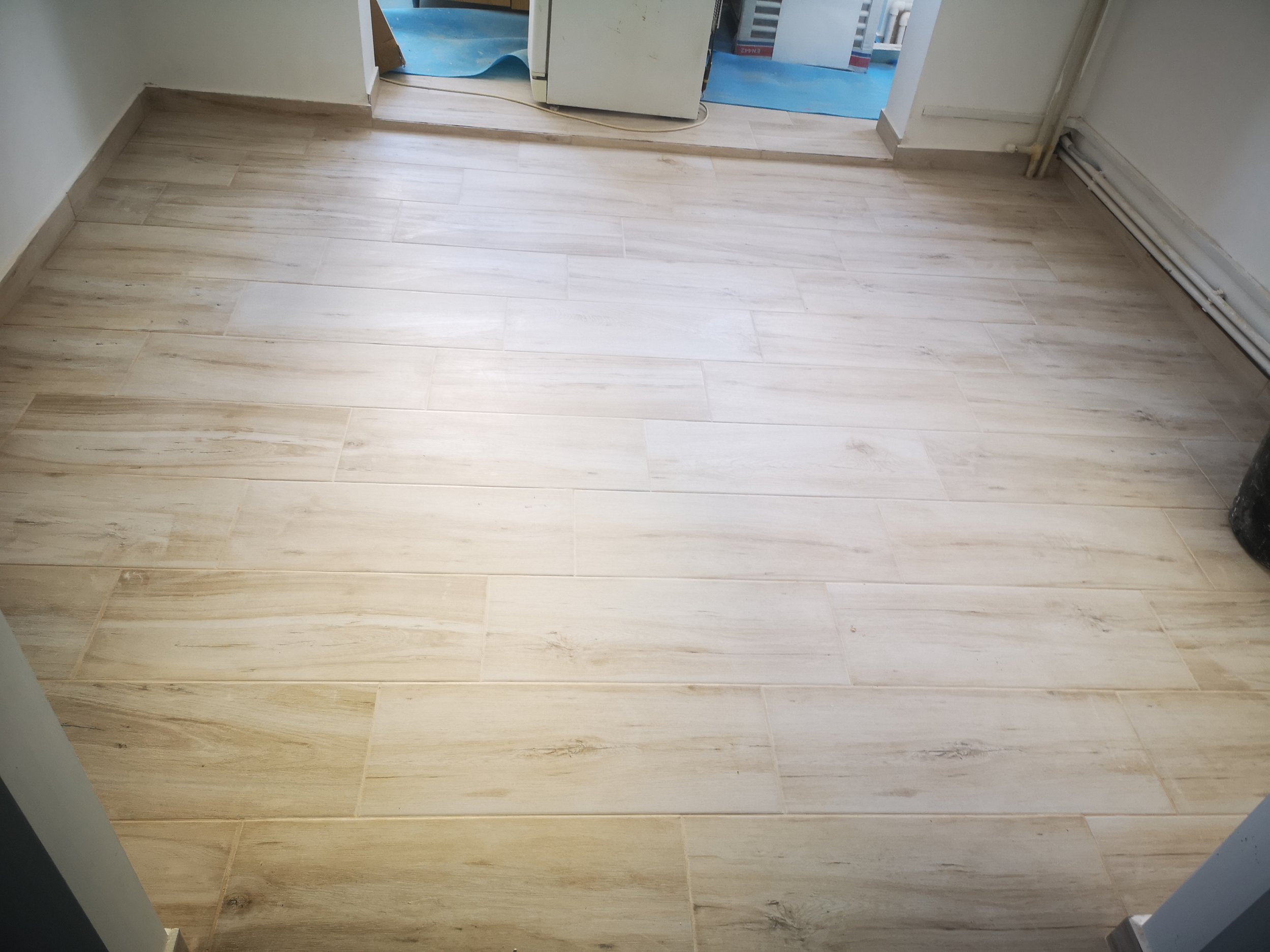 View of a room with wooden laminate flooring and white walls. There is a doorway at the back with an area under construction, showing blue protective covering on the floor and some building materials or equipment.