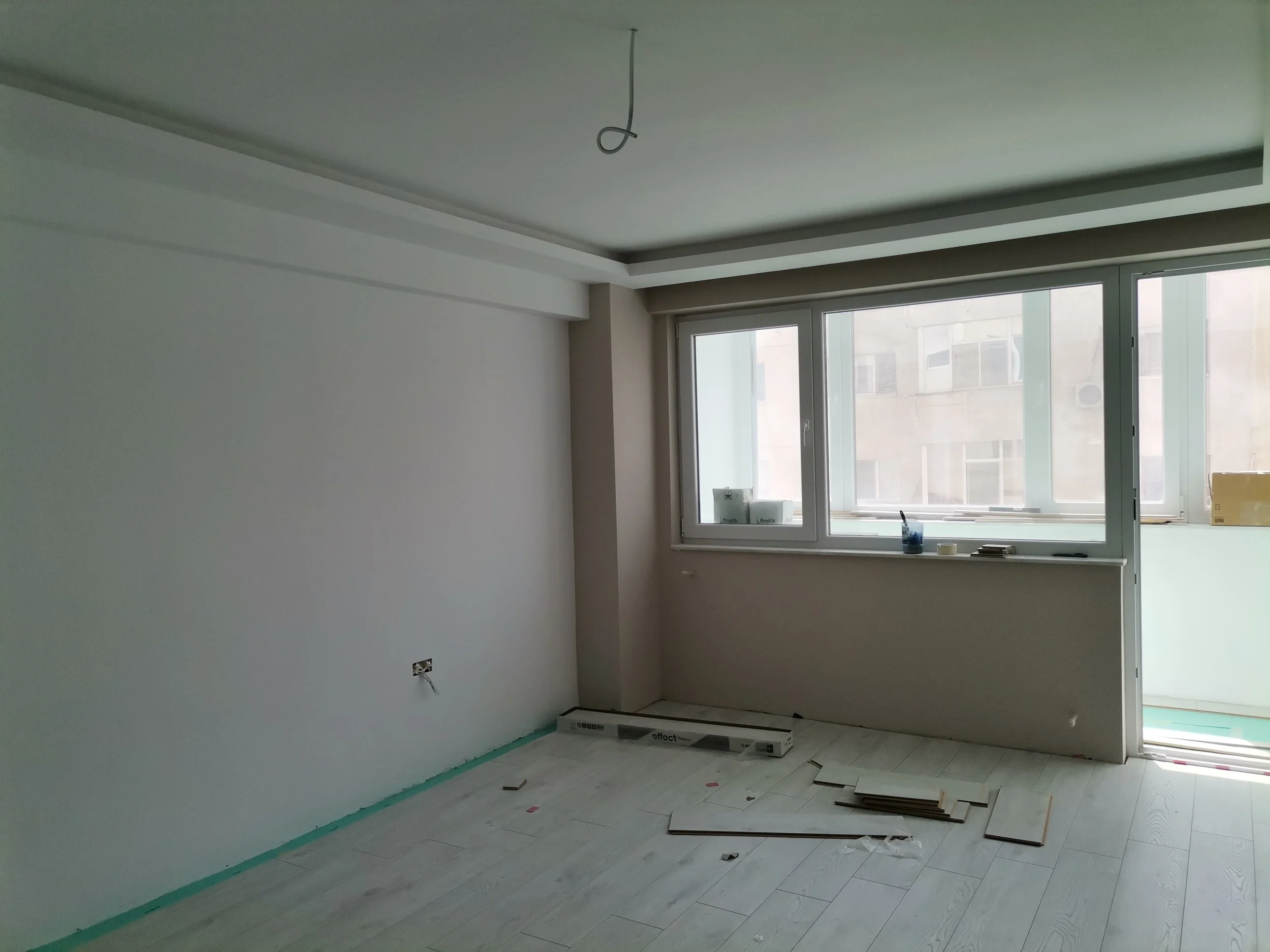 An unfinished room with light-colored wooden flooring, white walls, and large window with a view of neighboring buildings. Construction materials and tools are scattered on the floor, and a power outlet is visible on the wall.
