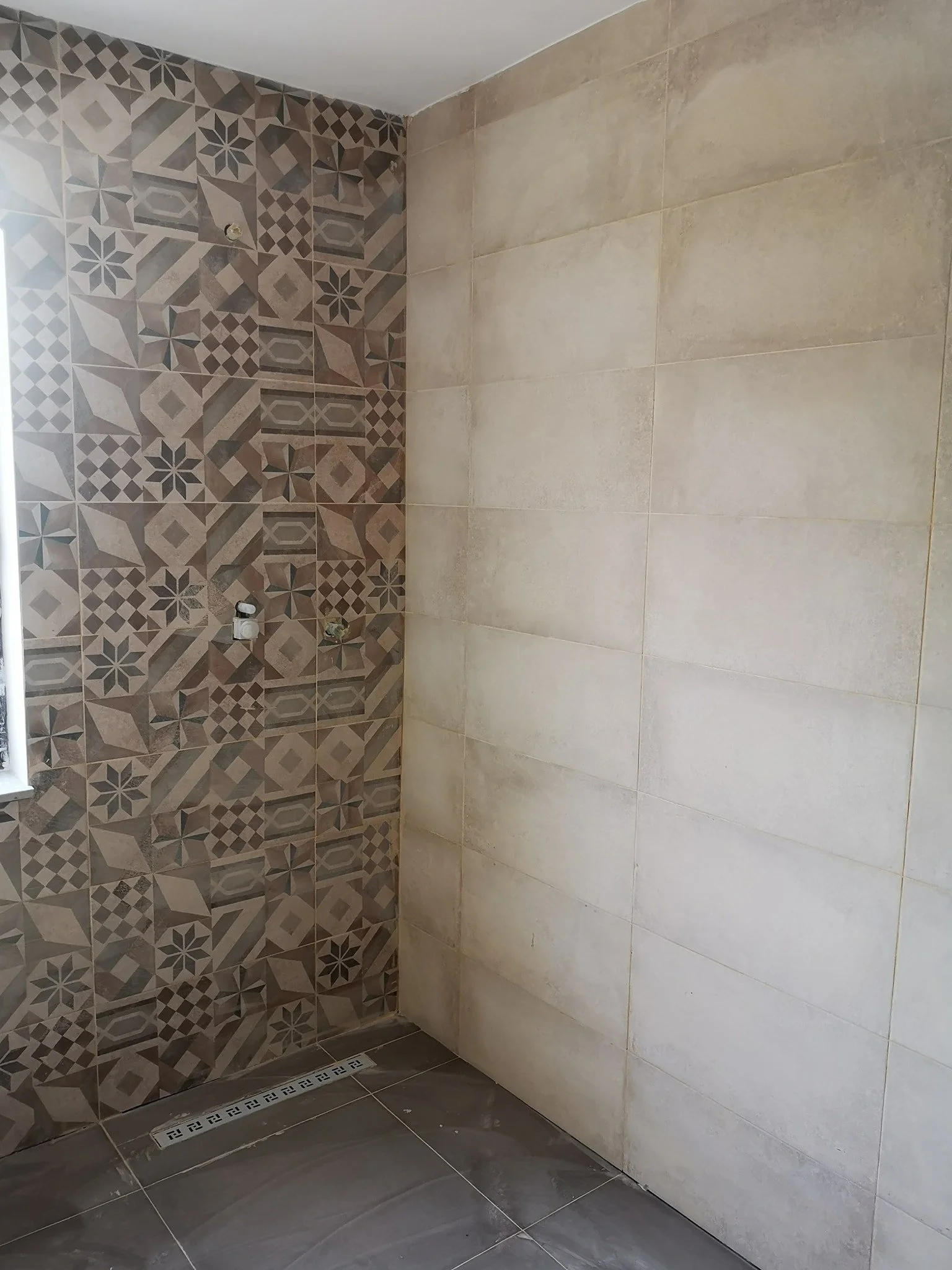 Bathroom shower area with decorative patterned tiles on the left wall and plain light-colored tiles on the right wall, with a linear floor drain at the base.
