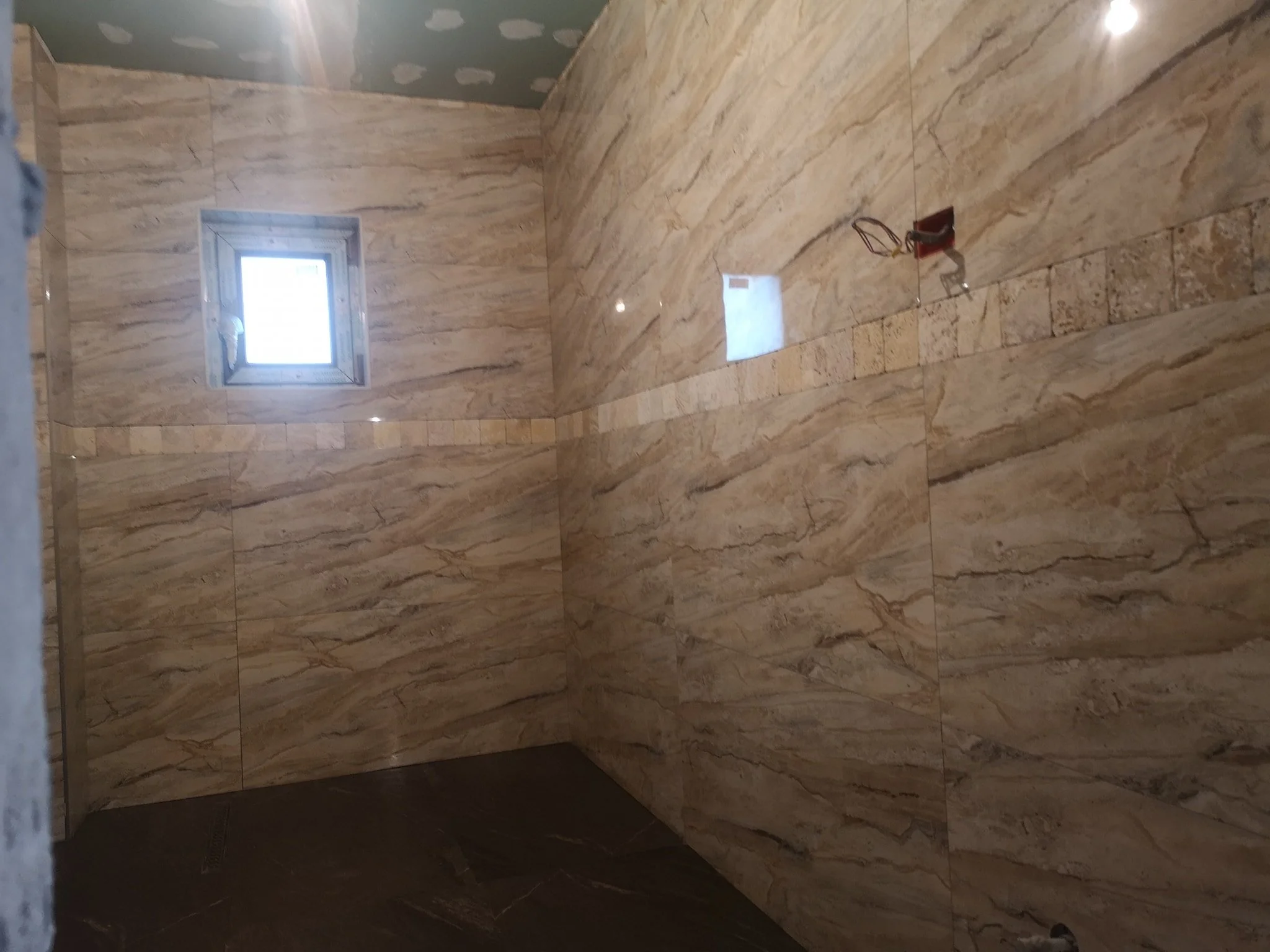 An unfinished bathroom corner with beige marble tile walls and a small window, showing electrical wiring and fixtures for a shower installation.