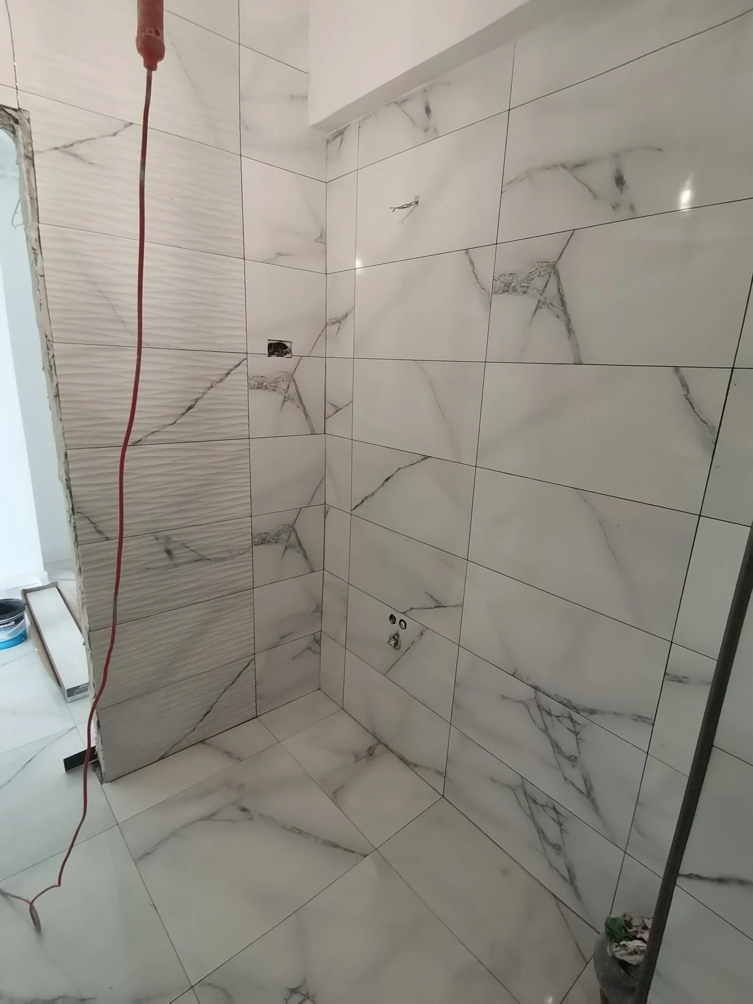 A bathroom under construction with large white marble tiles with gray veining on the walls and floor. There is a red cable hanging from the upper left, an unfinished electrical outlet on the wall, and a black basket with construction debris at the bo