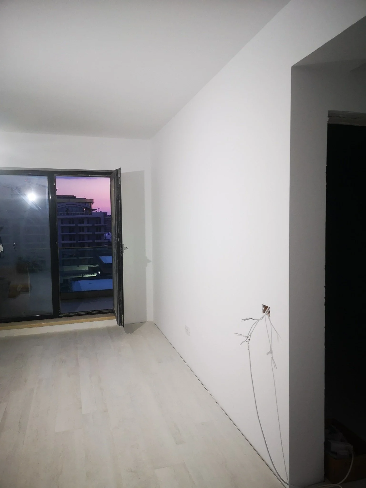 An empty room with white walls, a partially open window showing buildings and a purple sky at sunset, with some exposed electrical wires near the bottom right corner.