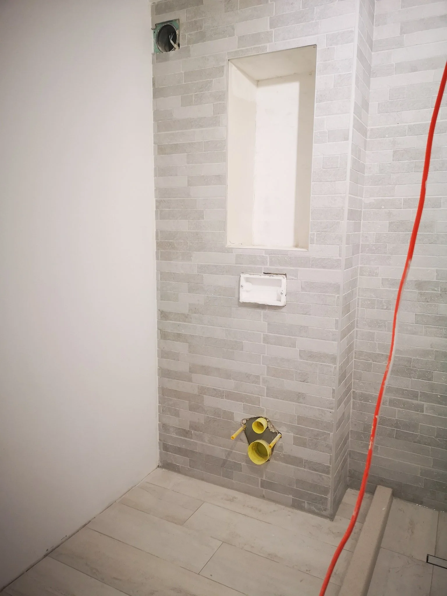 Bathroom under renovation with light-colored floor tiles, a partially tiled wall with gray brick-patterned tiles, and exposed plumbing pipes.
