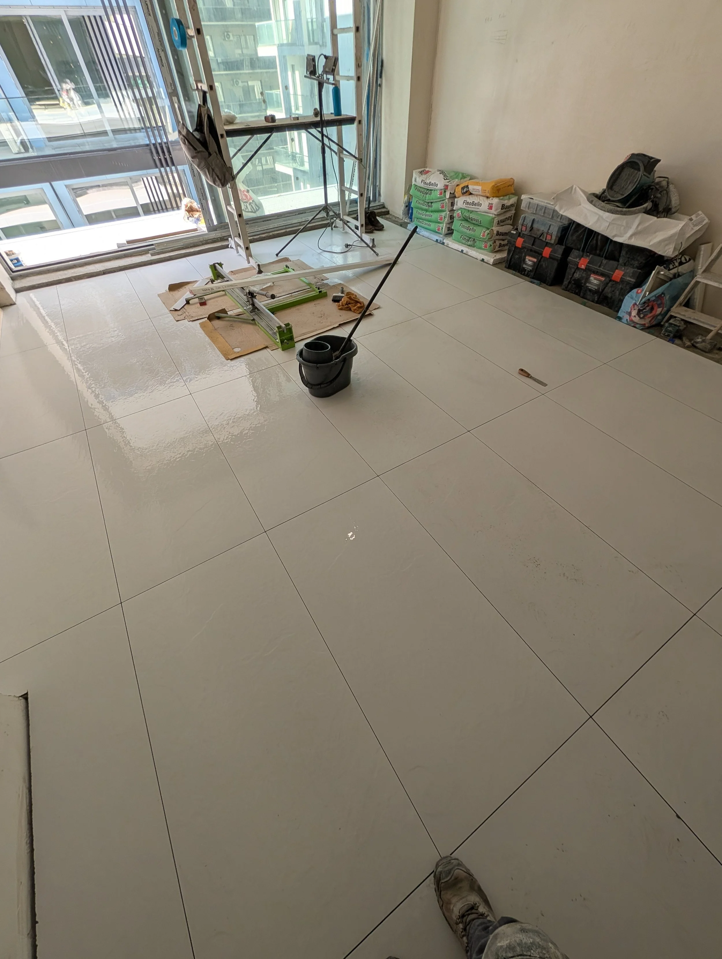 A person wearing dirty work shoes standing in a room with tiled floor, construction tools, and supplies. A window with a city view and a scaffold inside the room.