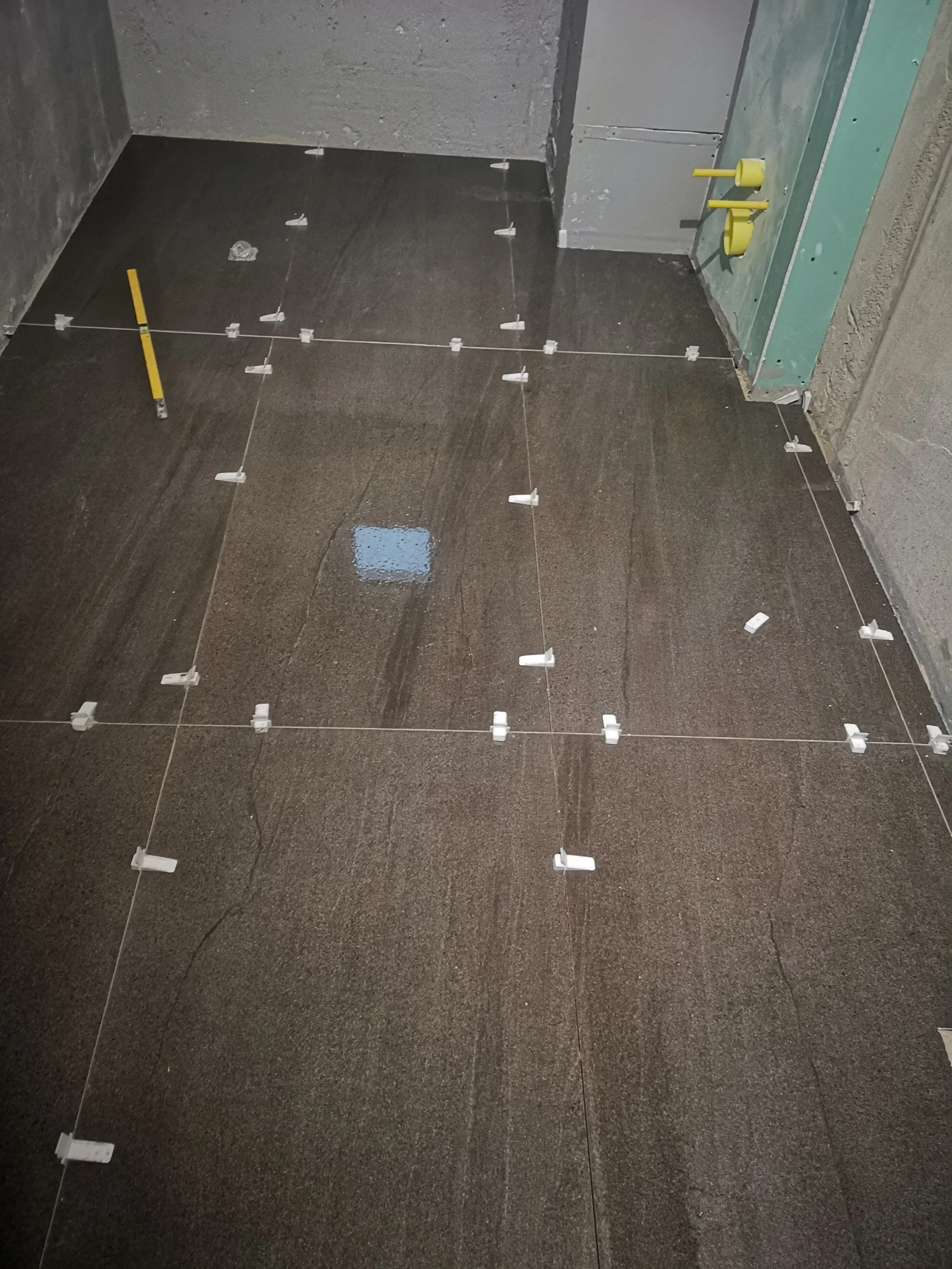 Construction site with a tiled floor, wall, and partially installed electrical or plumbing fixtures, with some tools and materials present.