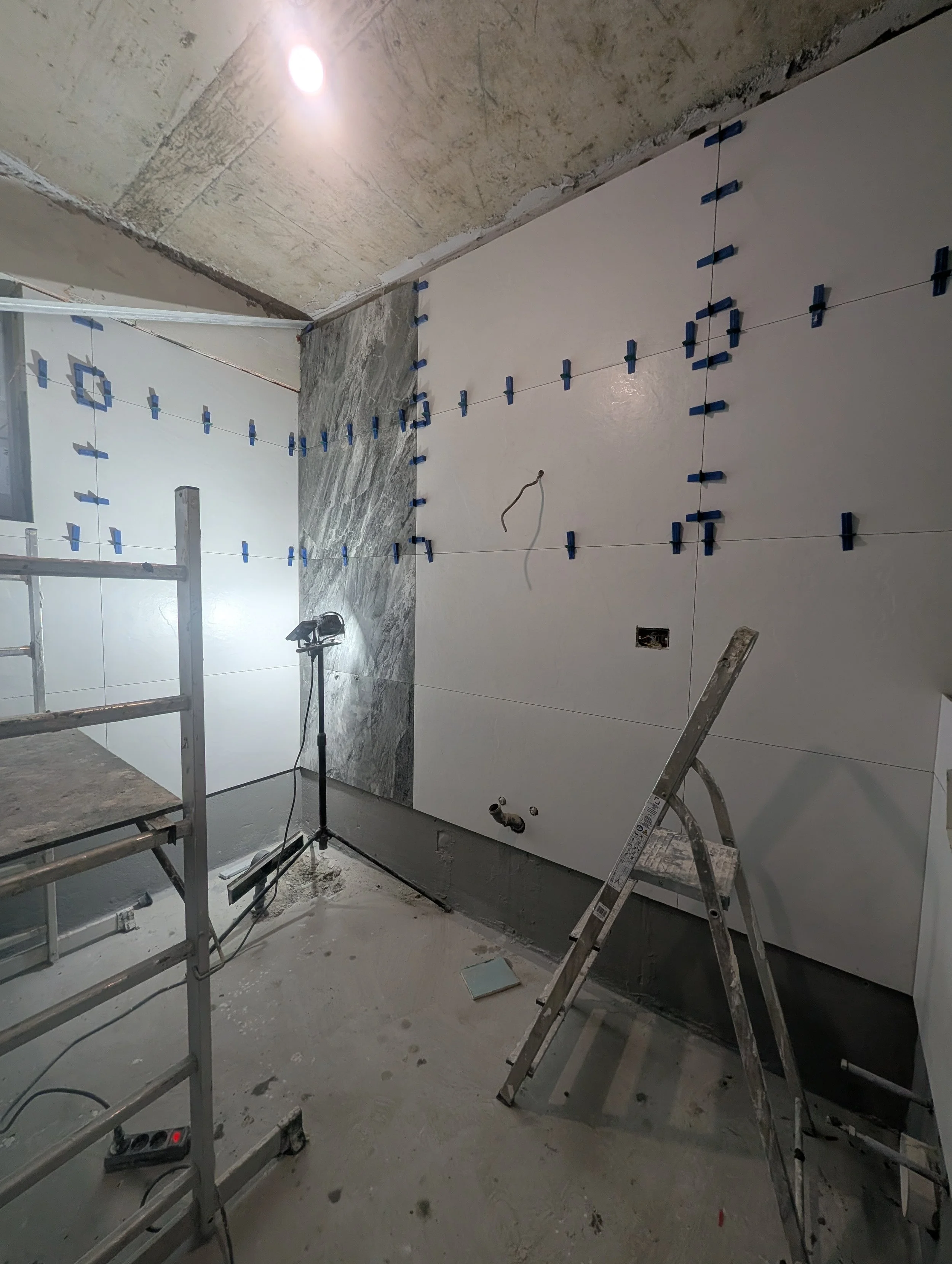 Bathroom under construction with large white wall tiles partially installed, construction tools, a ladder, scaffolding, and exposed ceiling.