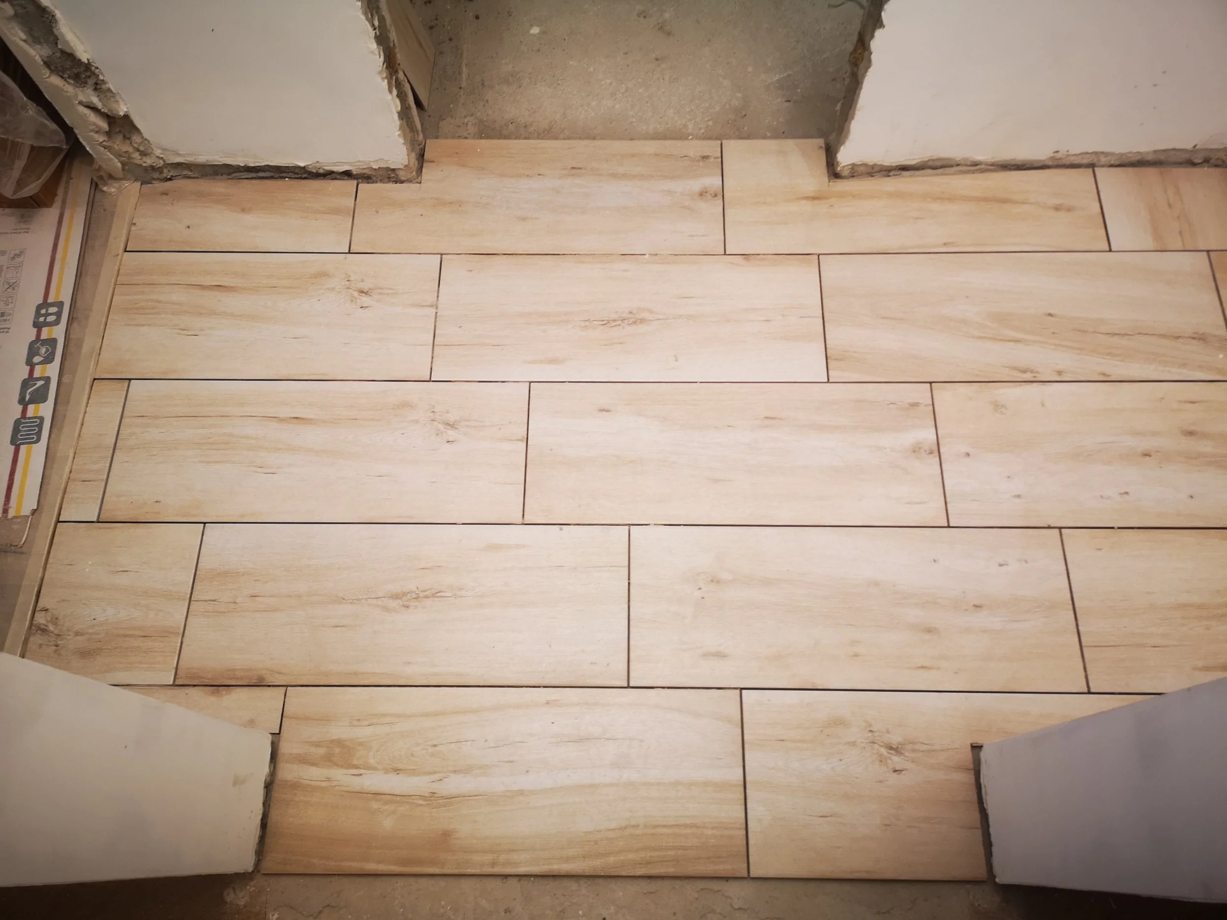 Tile flooring installation in progress in a room, with a section of the wall opening revealing the subfloor and unfinished wall area.