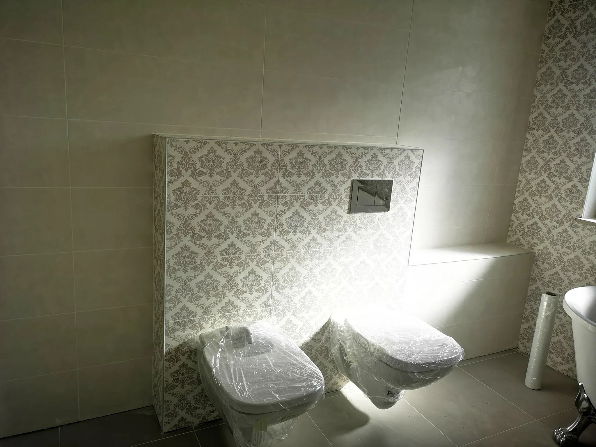 A bathroom with beige tile flooring and two toilets covered in plastic, a wall with beige decorative wallpaper, and a white toilet paper roll on a silver holder.