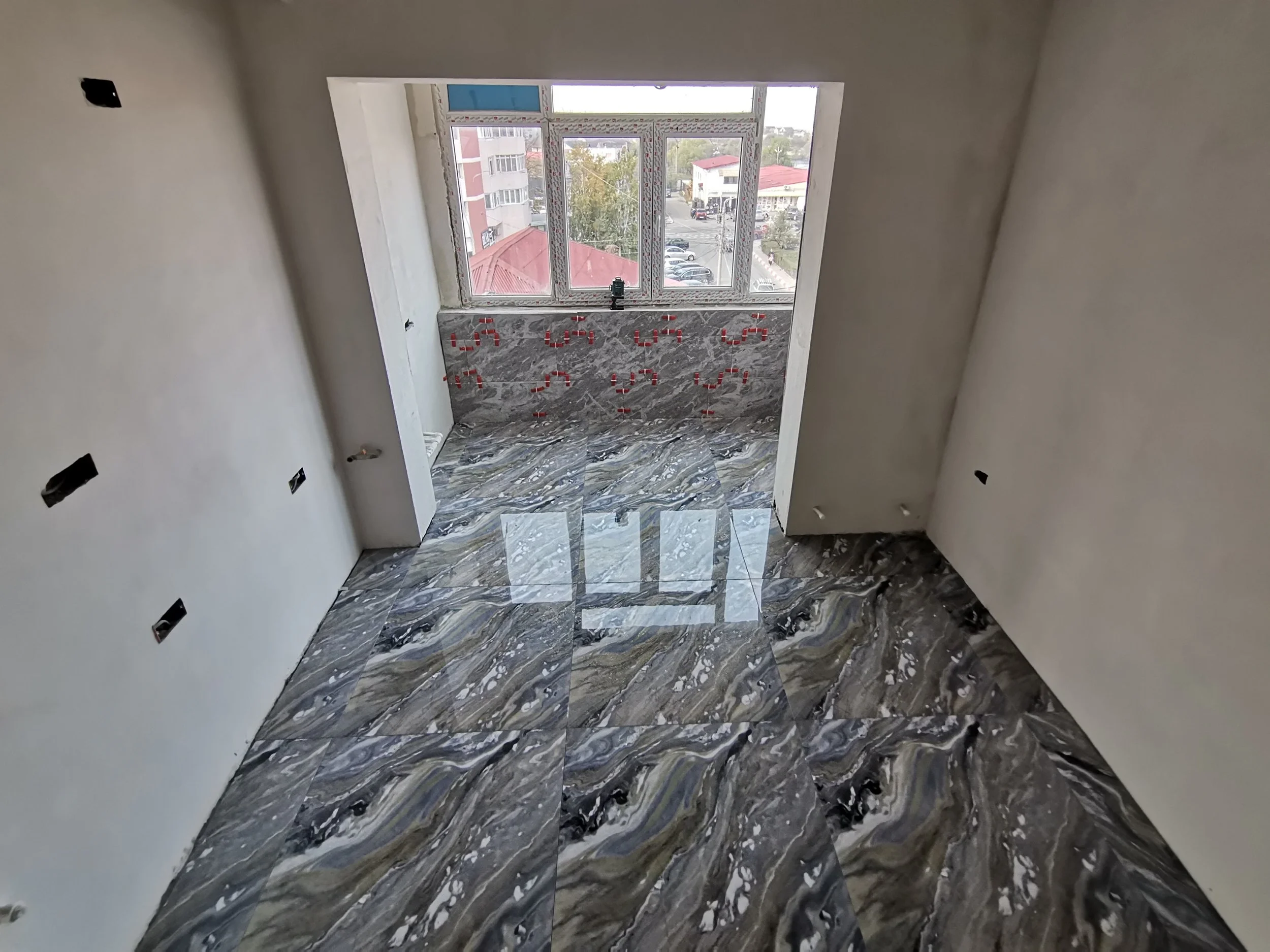 Interior of a room under construction or renovation with marble-like tiled flooring, a large window overlooking a street with buildings and parked cars, and unfinished walls with electrical outlets and wiring.
