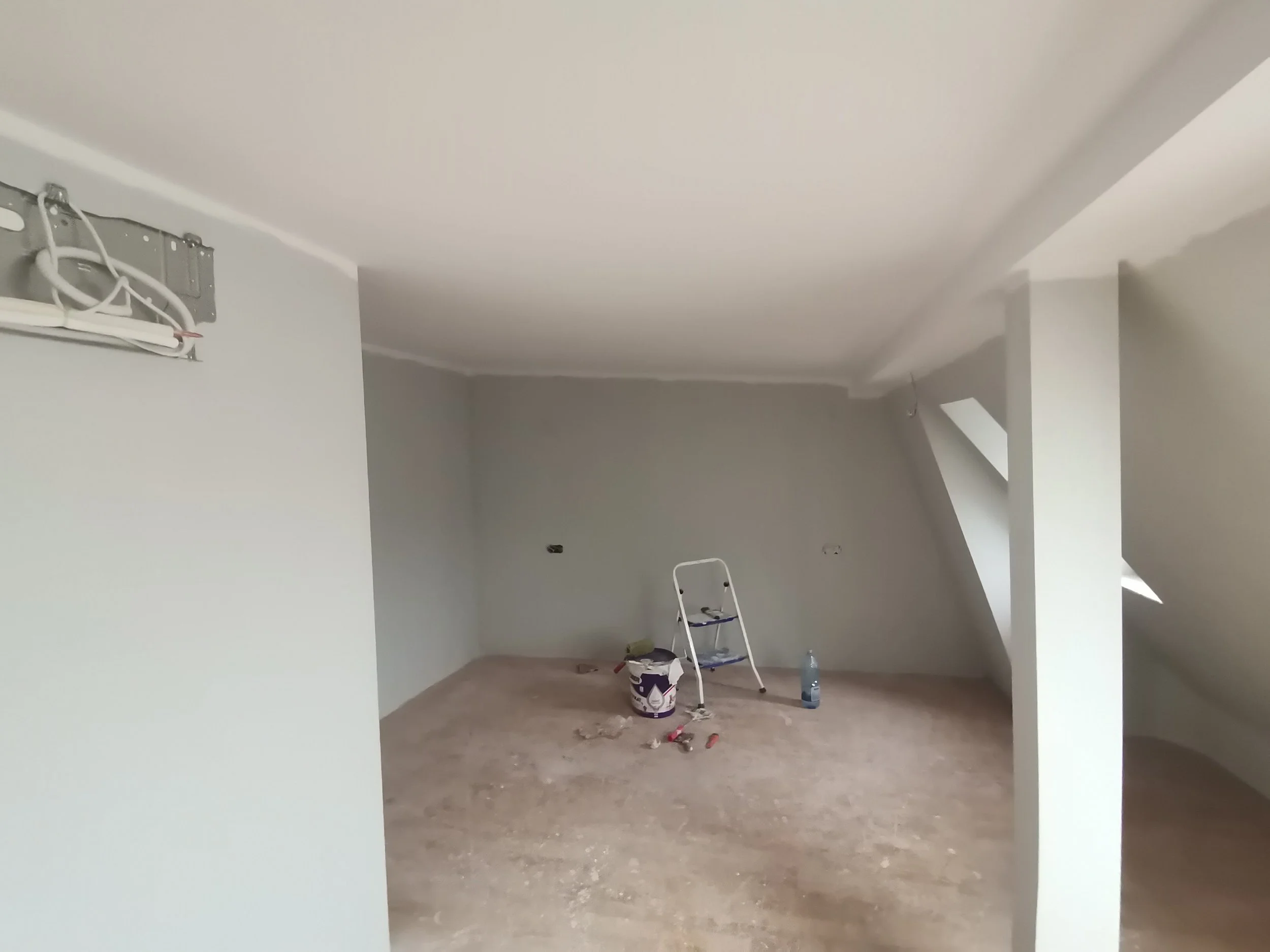 An unfinished room under renovation with a ladder, paint cans, a water bottle, and scattered tools on a bare floor, and electrical wires hanging from the ceiling.