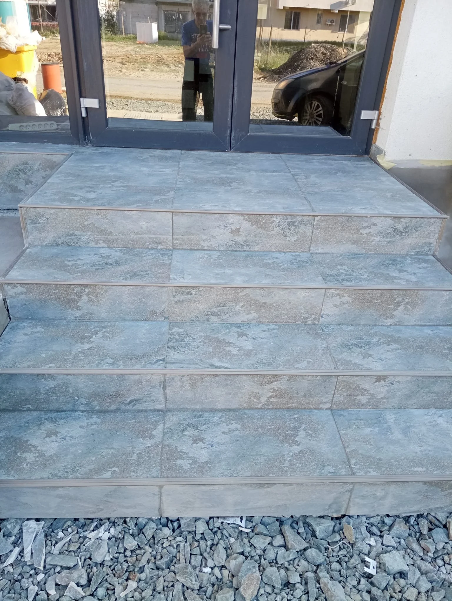 Glass door with stone steps leading up to it, and gravel at the base, with a person taking a photo reflected in the door.