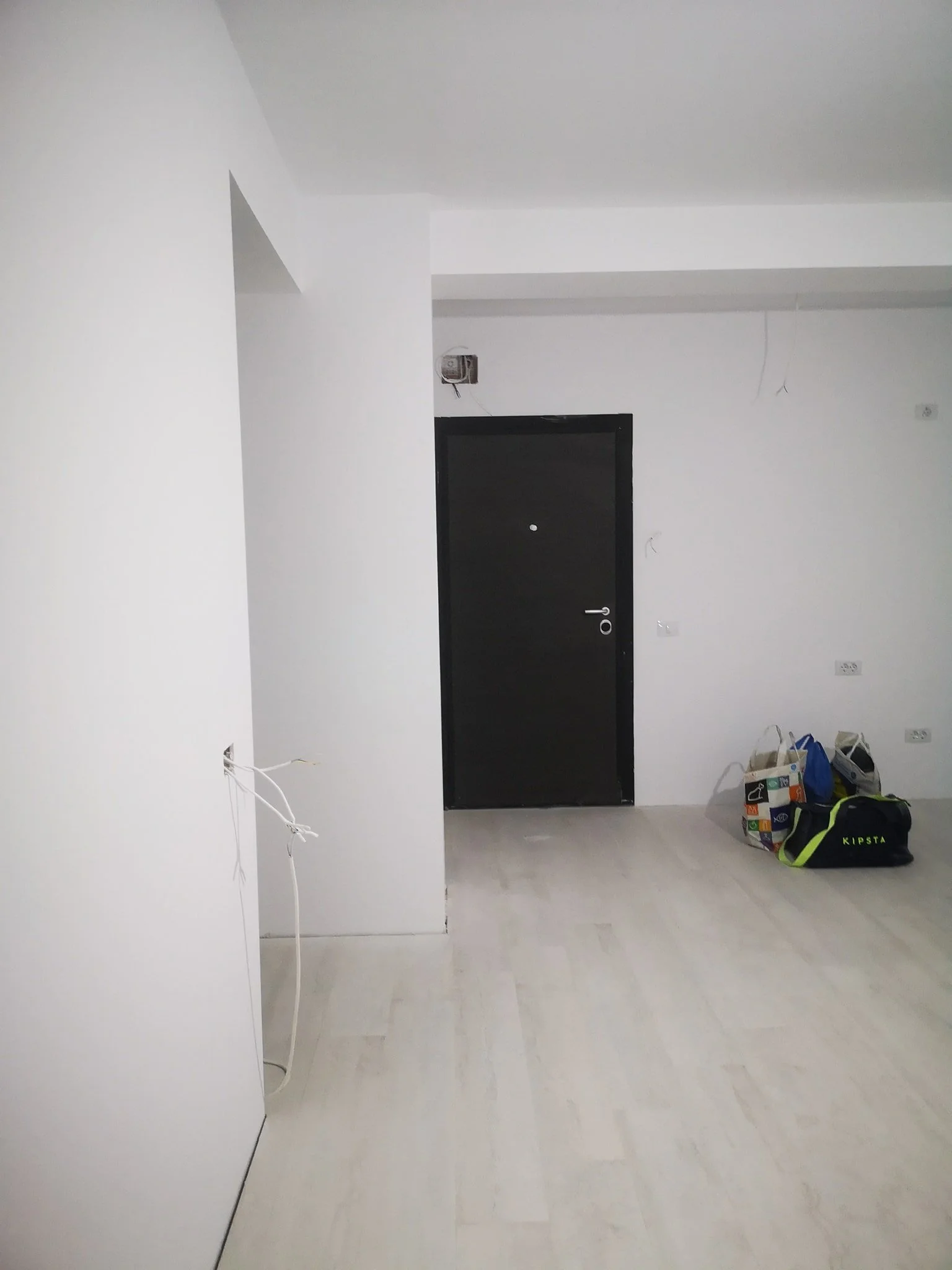 Empty room with white walls and light-colored flooring, some construction or renovation items including shopping bags and a duffel bag on the floor, black door, and visible electrical wiring and outlets.