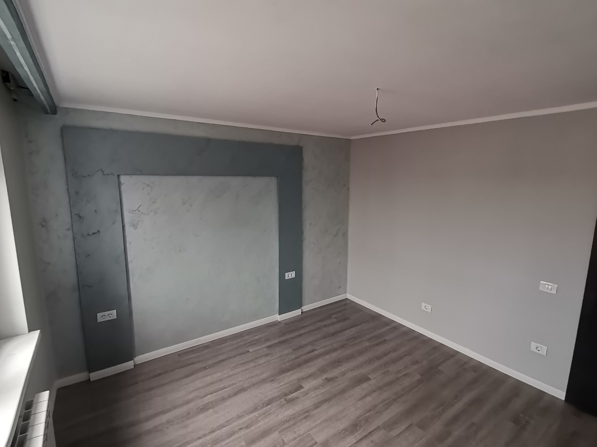 An empty room with partially painted walls, electrical outlets on the walls, and a ceiling with a wire hanging down. The floor is covered with dark wood laminate.