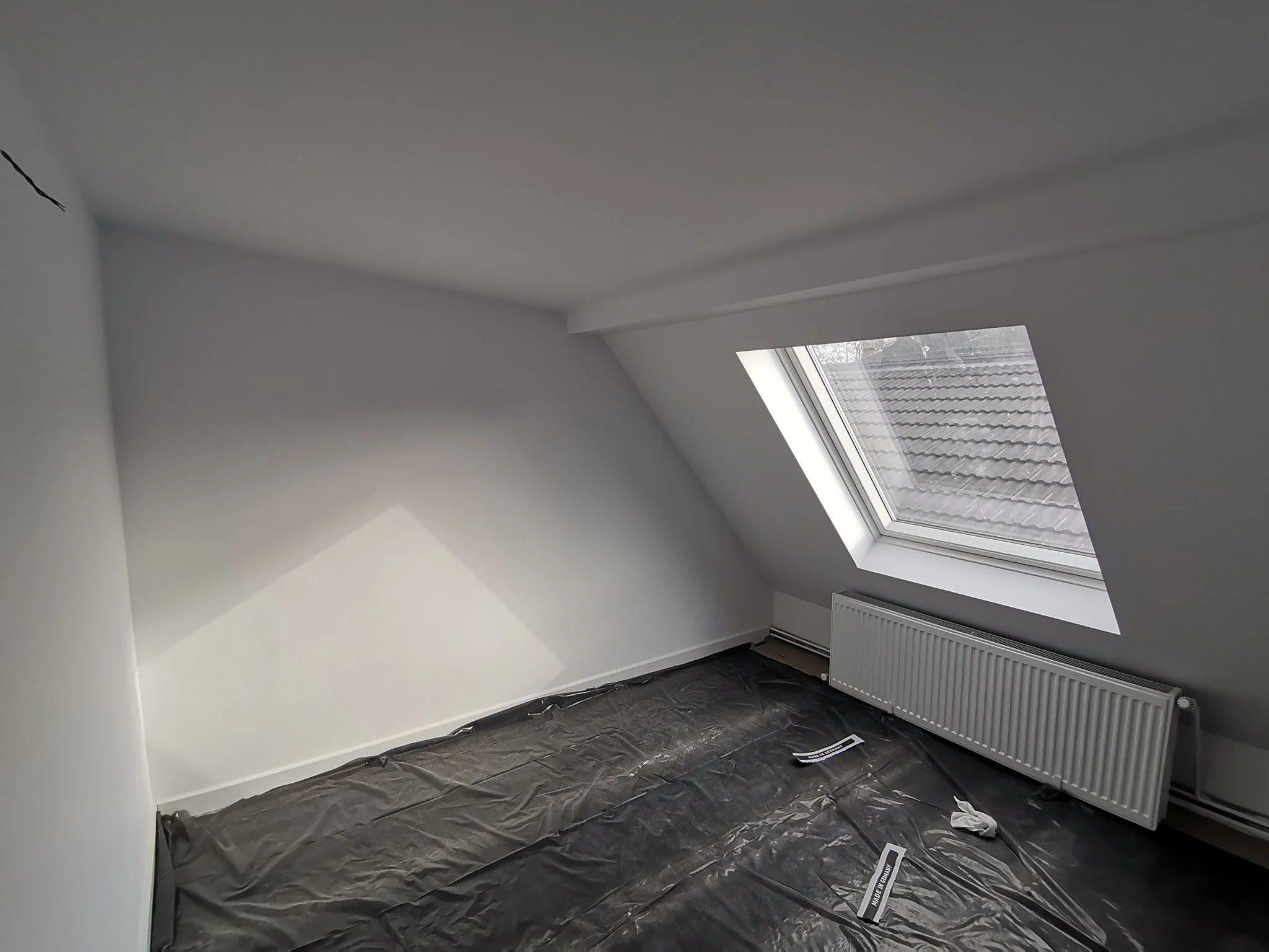 An empty attic room with white walls and a ceiling, featuring a large skylight window showing roof tiles outside, and a white radiator beneath the window. The floor is covered with black plastic sheeting.