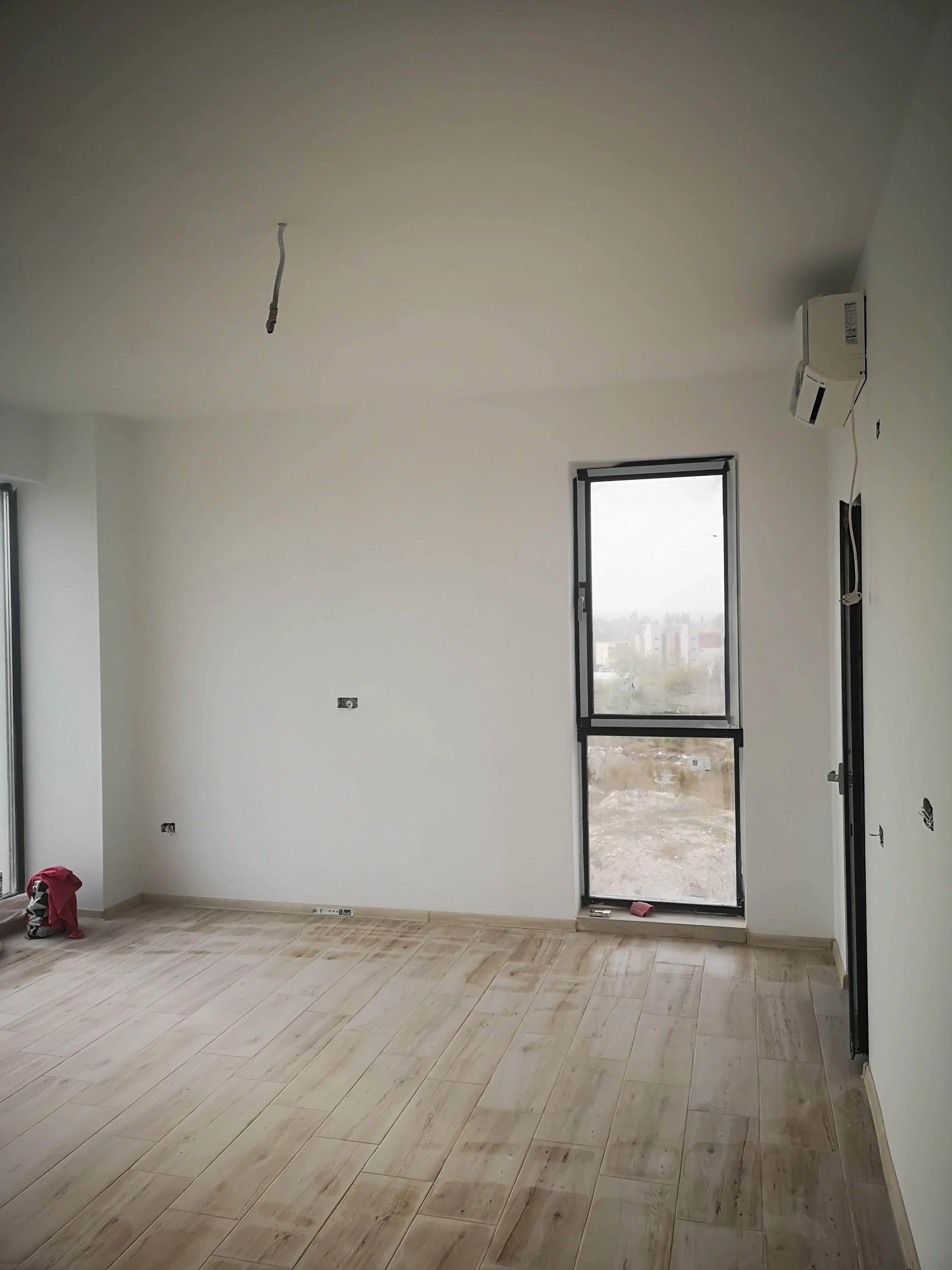Unfinished modern room with white walls, light wood flooring, a large window, and an air conditioning unit on the wall. Construction tools placed on the floor.