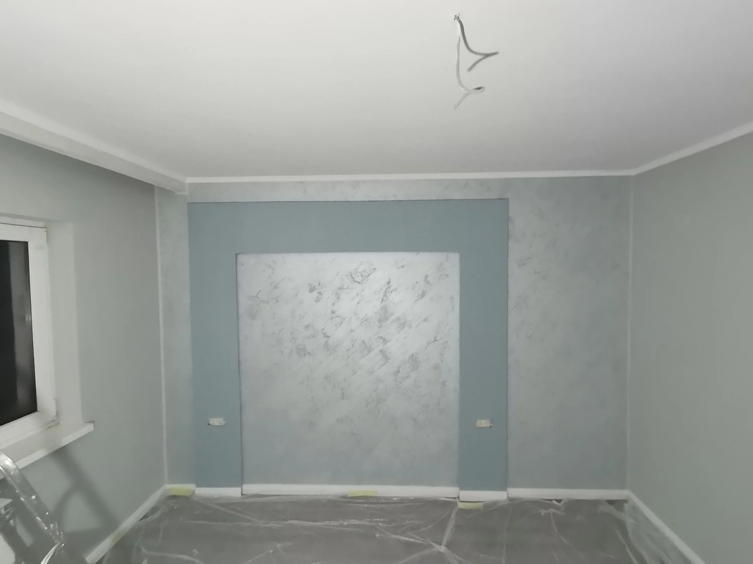 A room in the process of being painted with light blue walls, a white ceiling, and a window on the left side. Painting supplies are covered with plastic on the floor.