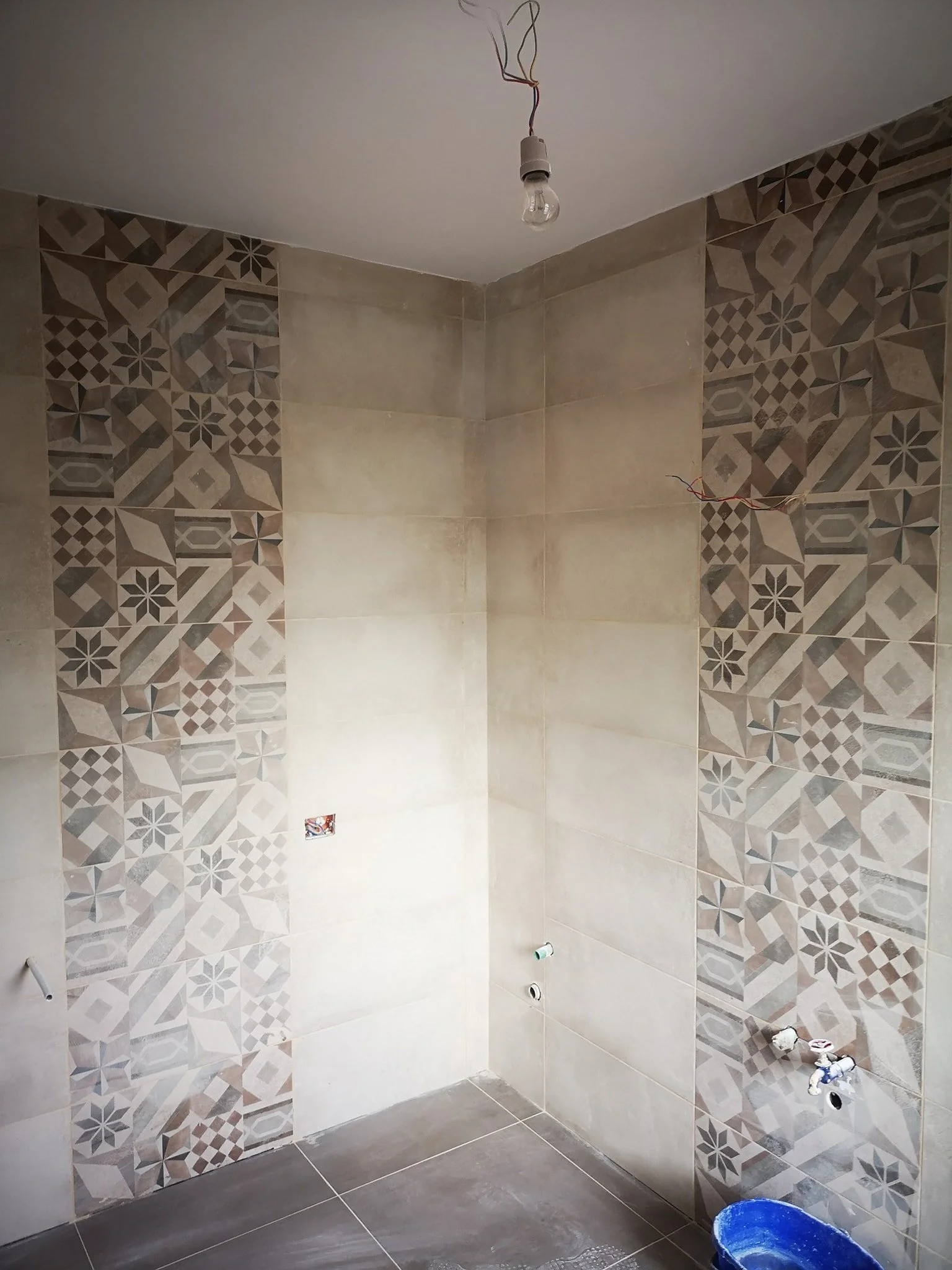 A partially tiled bathroom wall with decorative patterned tiles on either side, a white ceiling with exposed wiring and a light bulb socket, and plumbing fixtures near the floor.