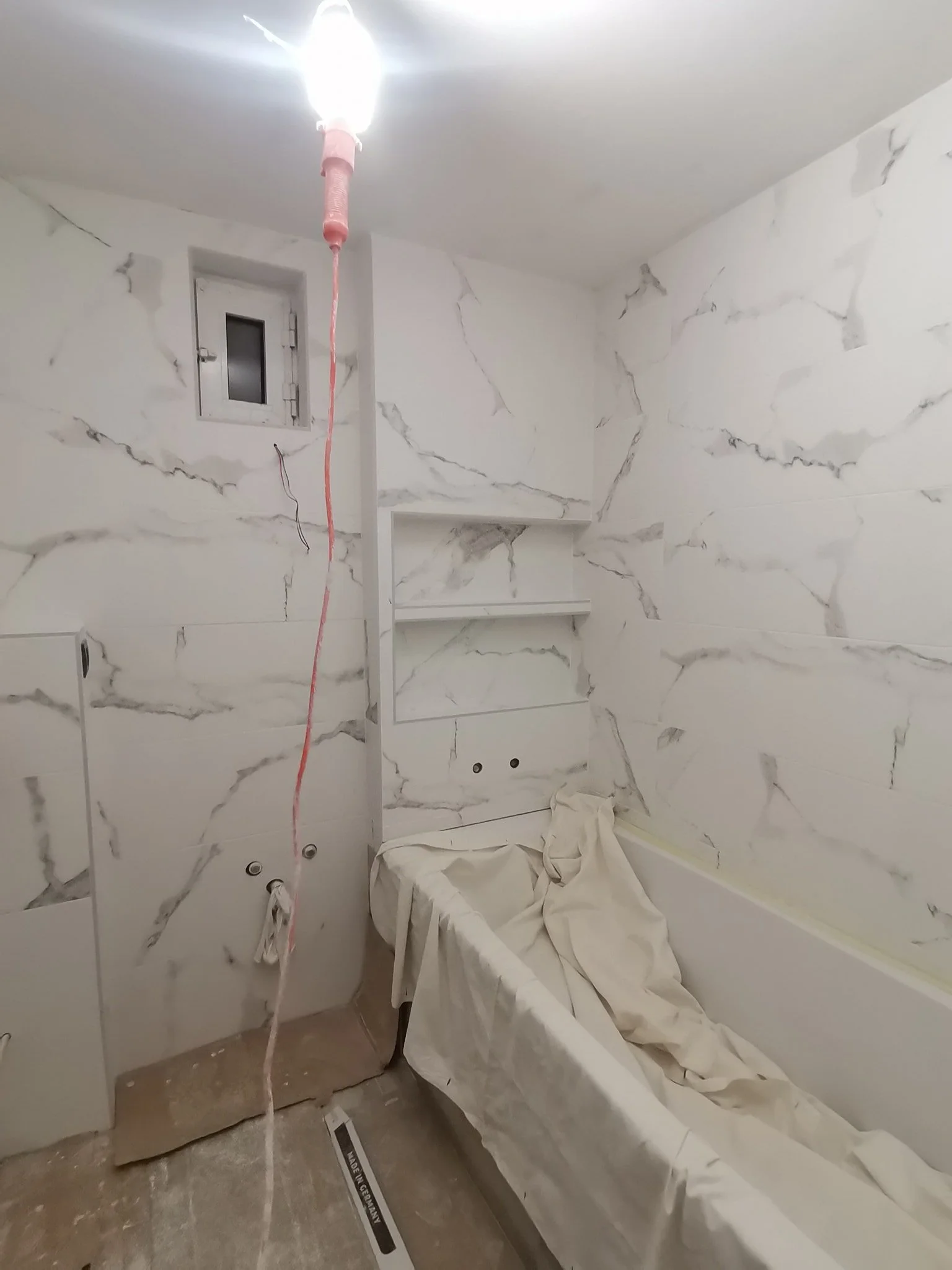 Bathroom with marble-patterned wall tiles that are still being installed, construction tools and materials, a small window, and protective coverings on some surfaces.