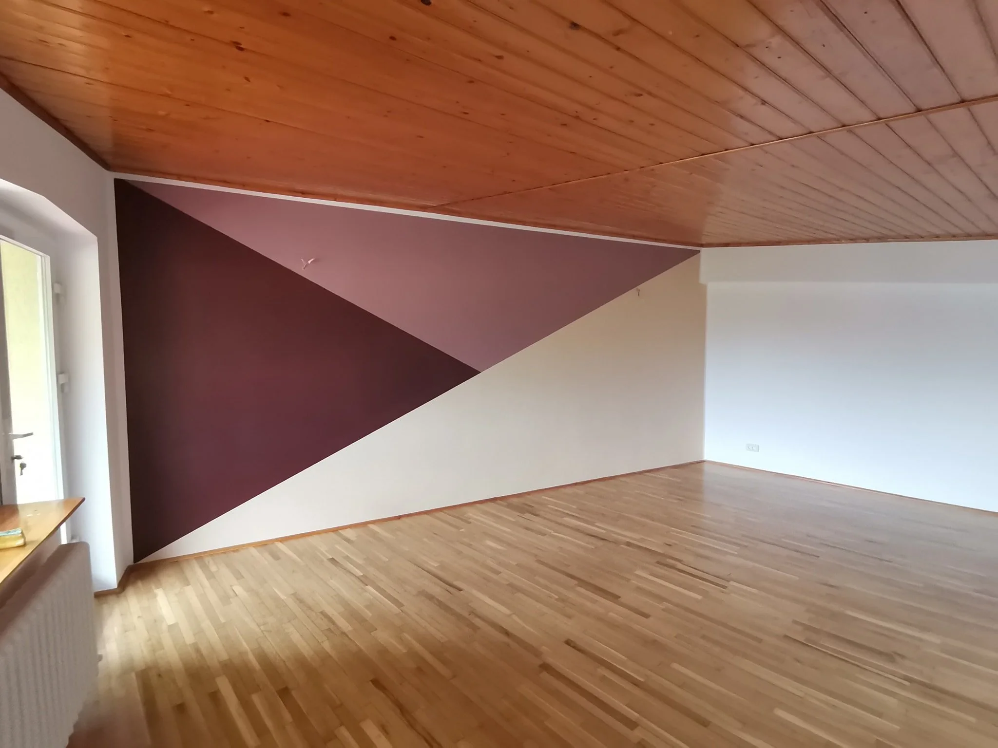 Empty room with wooden floor and ceiling, white and burgundy painted walls, and a window on the left side.