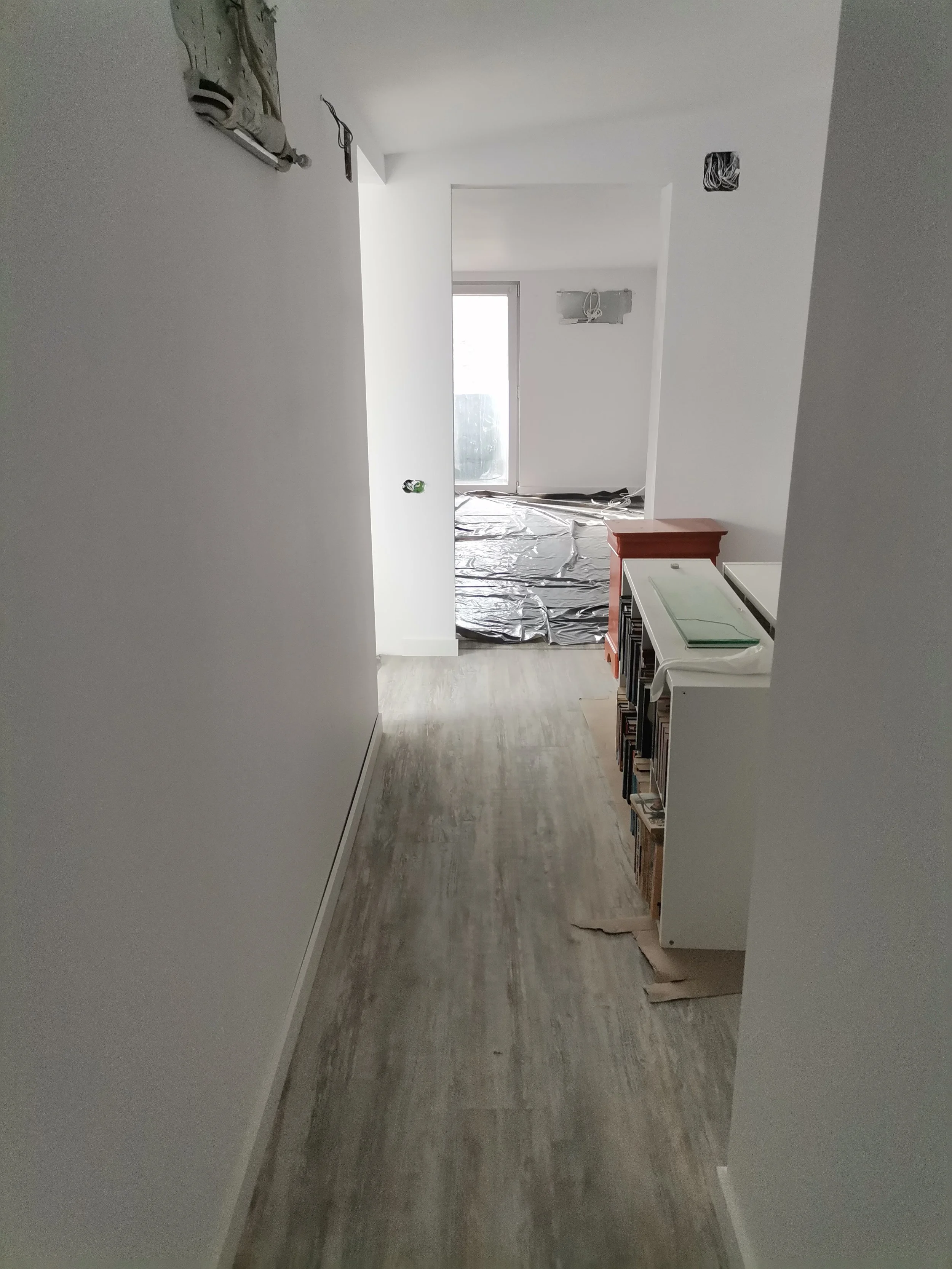 View down a narrow hallway in a home under renovation, with white walls, a wooden floor, and exposed electrical wiring. At the end of the hallway is a sliding glass door leading to an outdoor area, with plastic sheets on the floor and cardboard and t