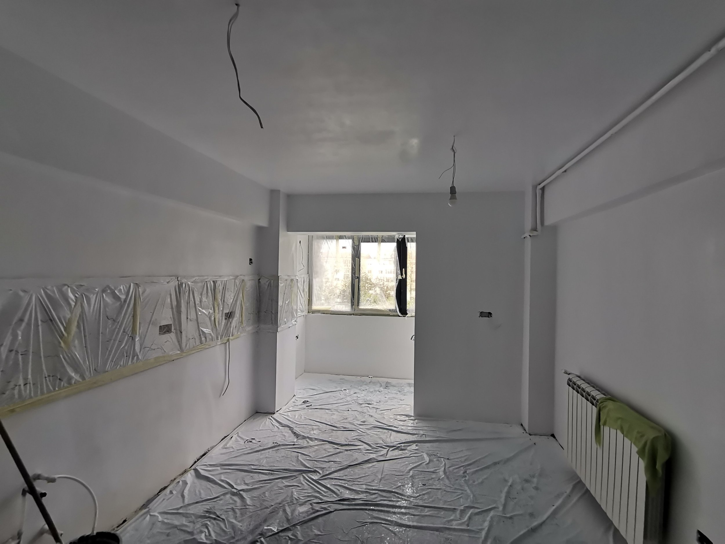 An indoor room under renovation with plastic sheets on the floor, hanging wires from the ceiling, and a window at the far end.