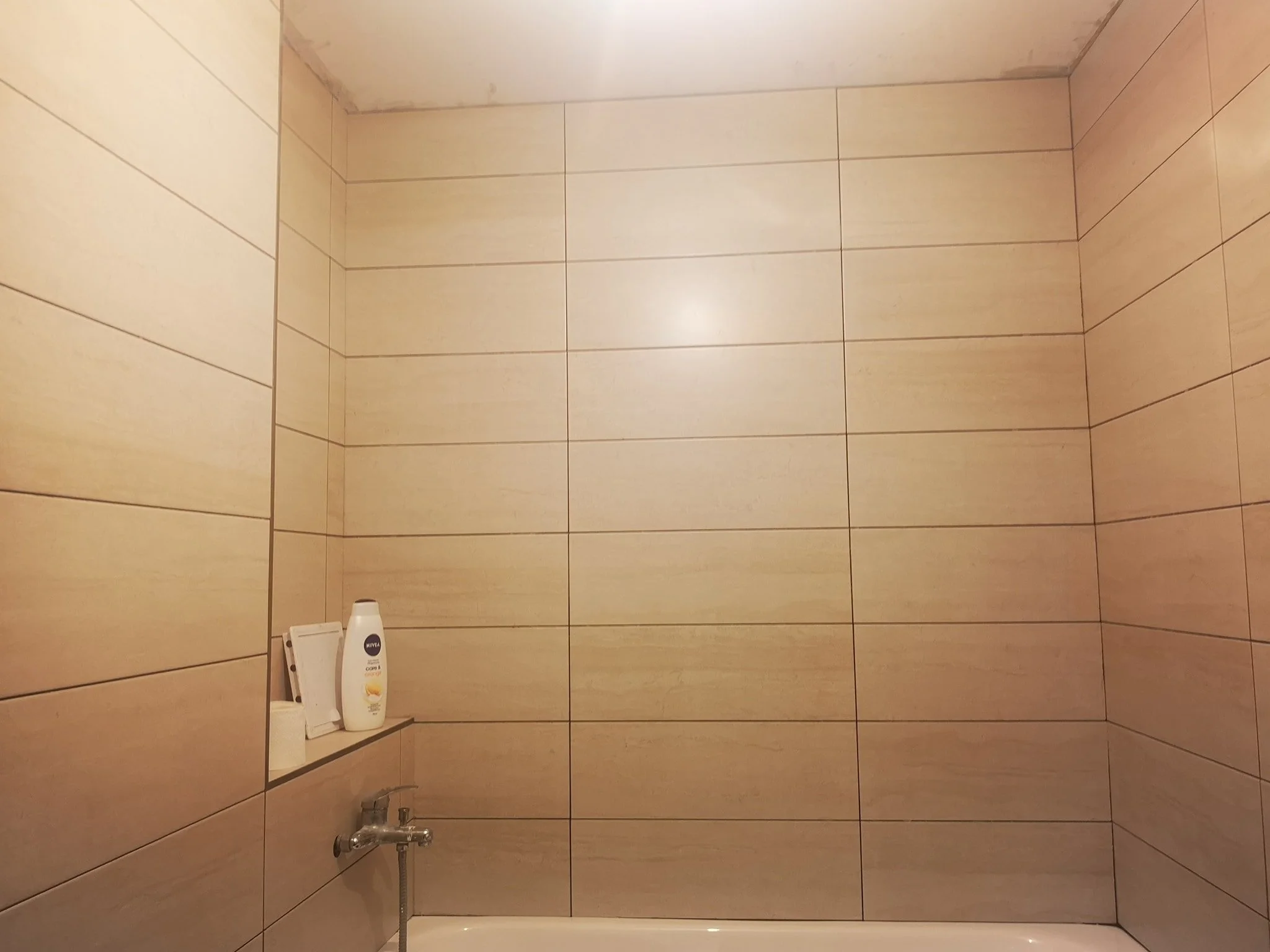 A shower area with tiled walls, containing a bottle of Nivea lotion, a white phone, and a small mirror.