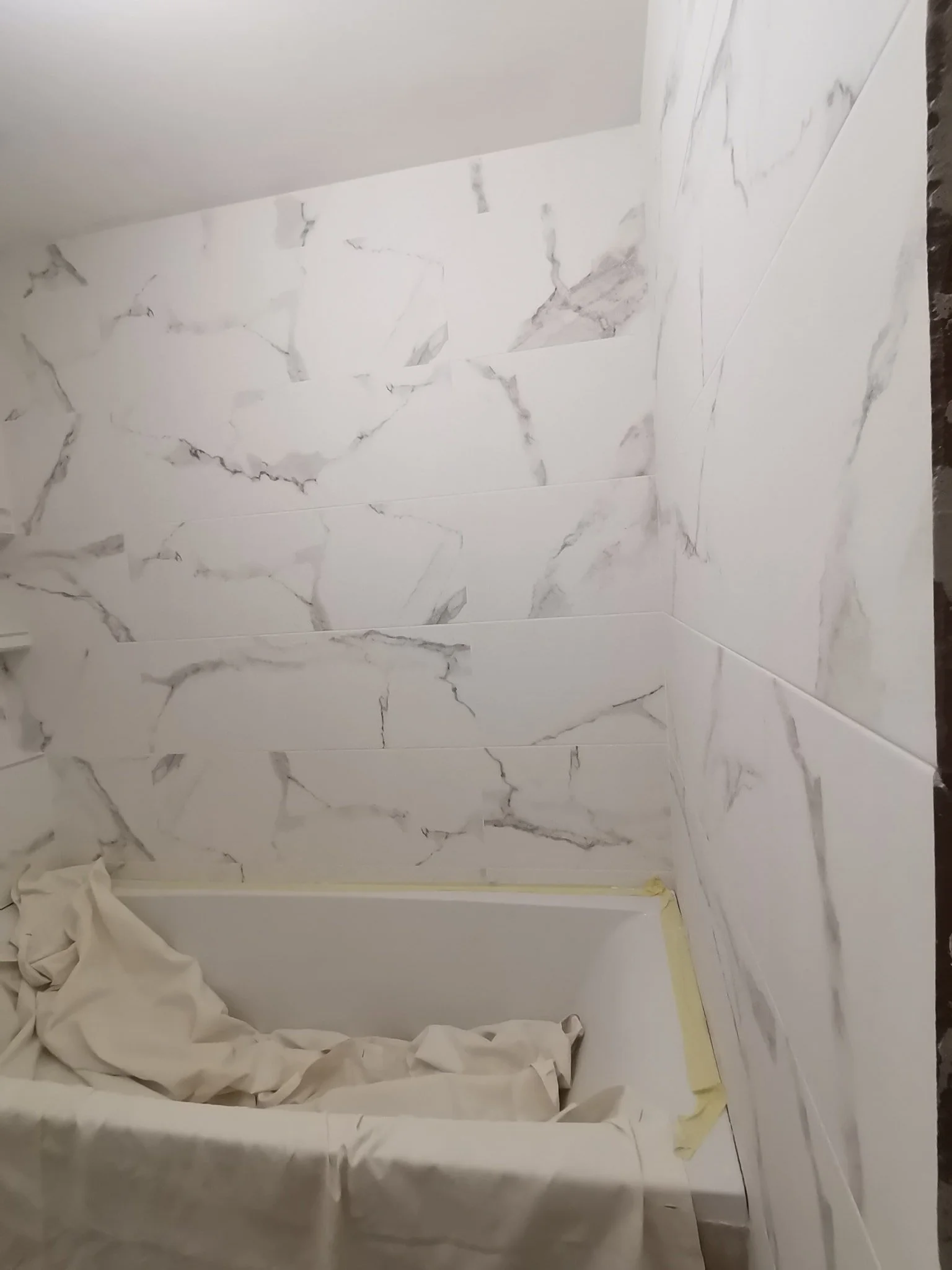 Bathroom walls and floor being tiled with white marble-look tiles, some tiles still need grouting, and painter's tape is visible along the edges.