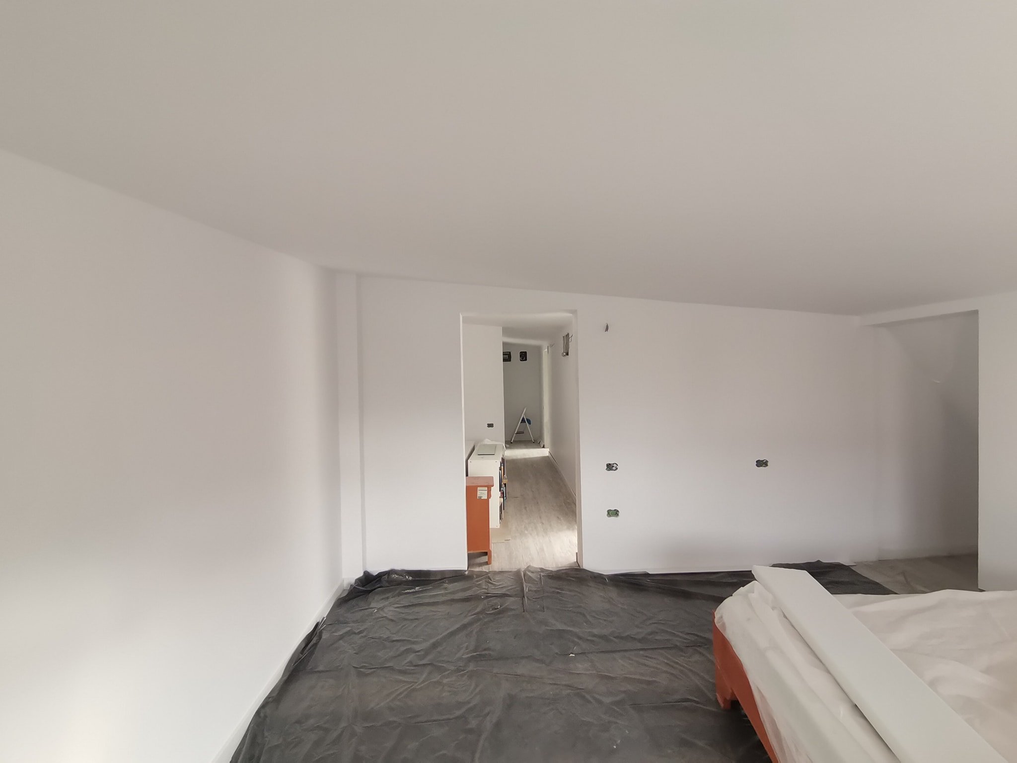 Interior of a room under construction with white walls and ceiling, plastic covering on the floor, and some construction materials and tools visible.
