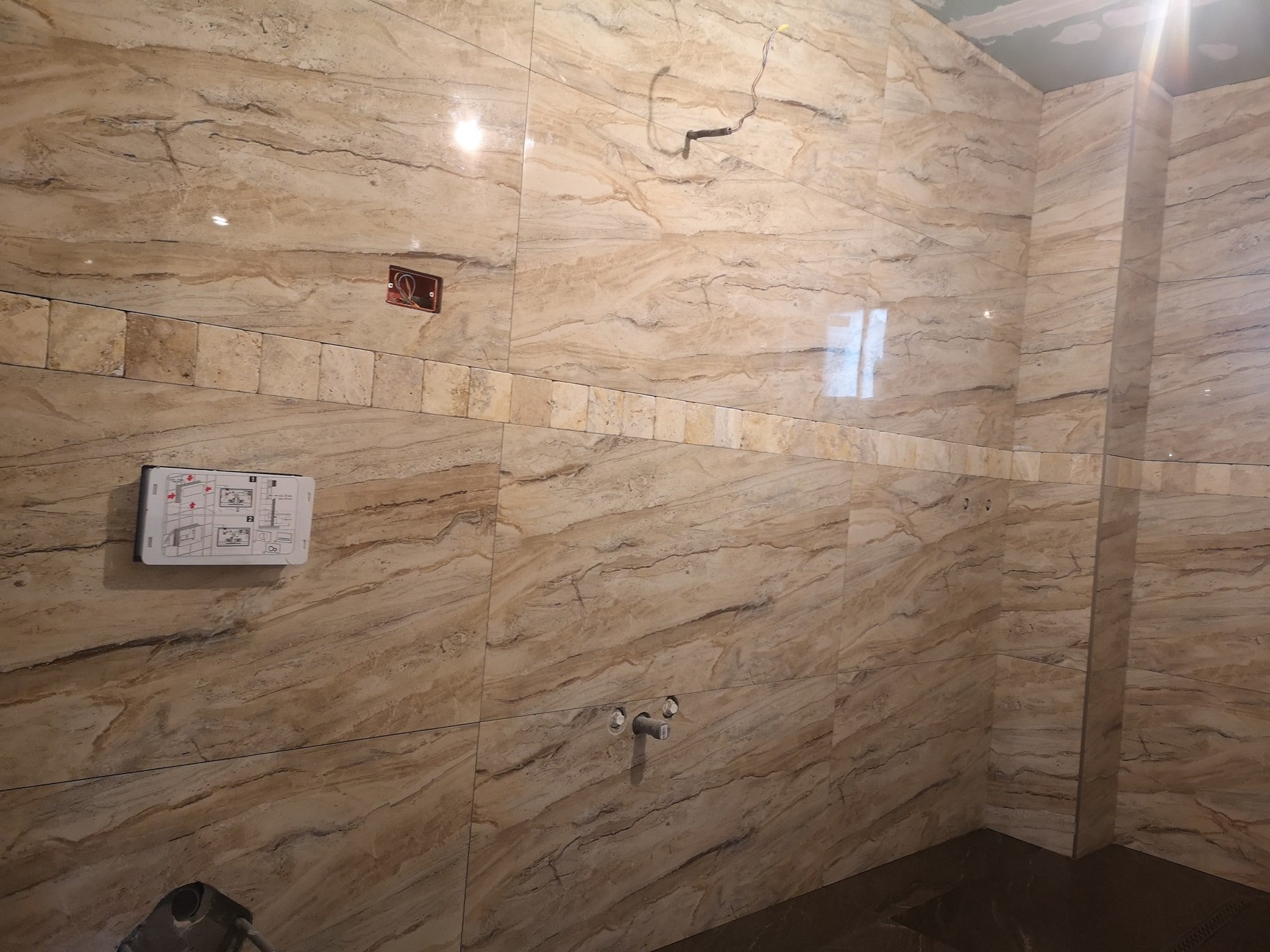 Bathroom wall with beige marble tile walls, exposed wiring, electrical outlet, plumbing pipes, and safety equipment.