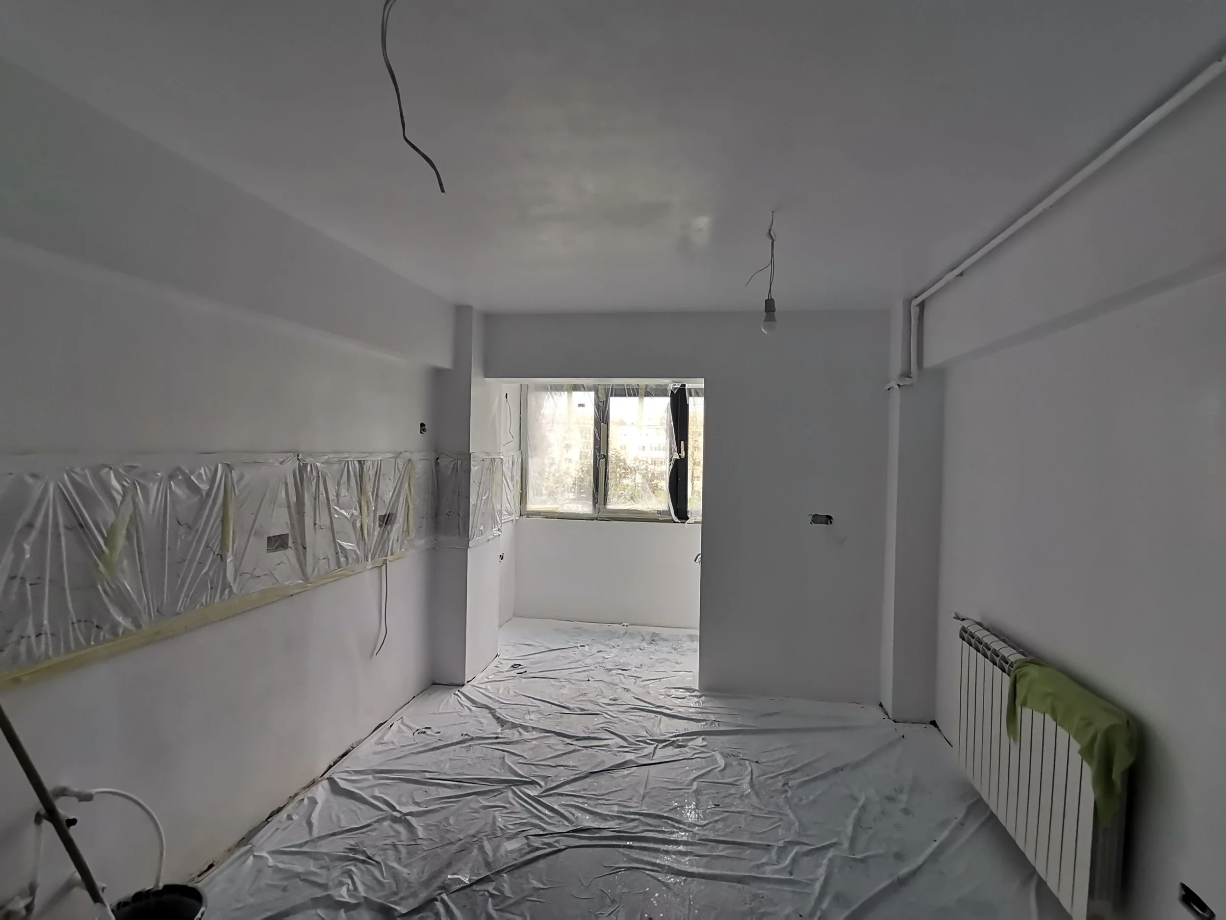 Interior of a room under renovation with plastic covered floor and walls, exposed wires hanging from the ceiling, and a window at the far end letting in natural light.