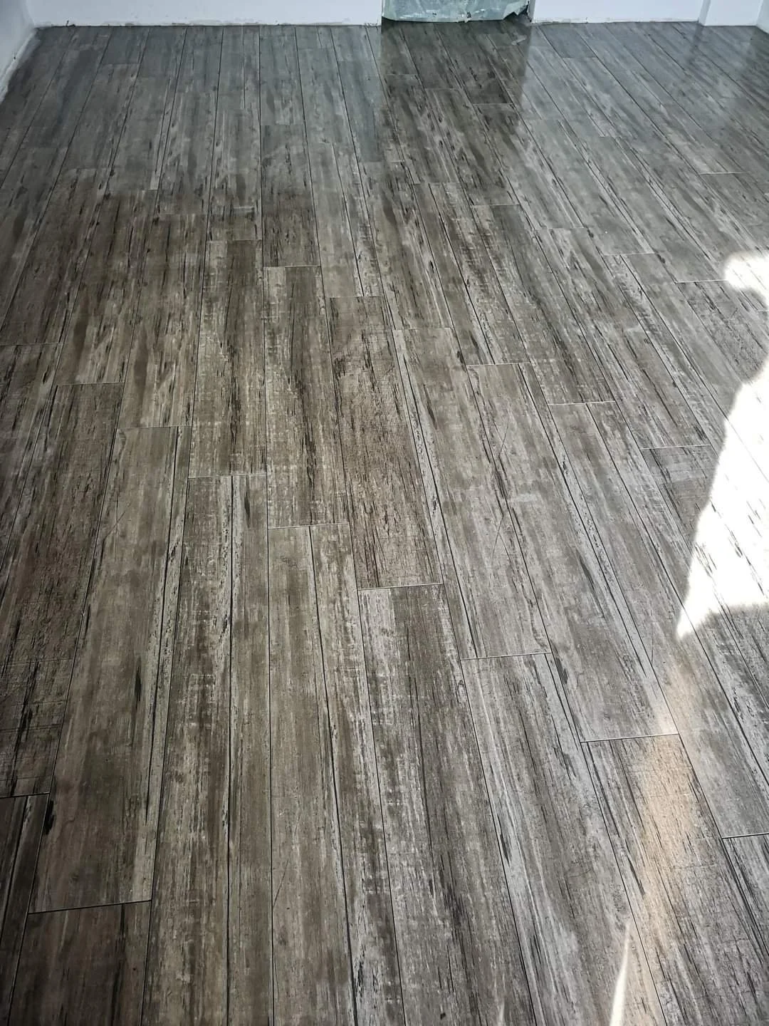 Wood-look tile flooring with gray and brown tones in a room with white walls and a corner.