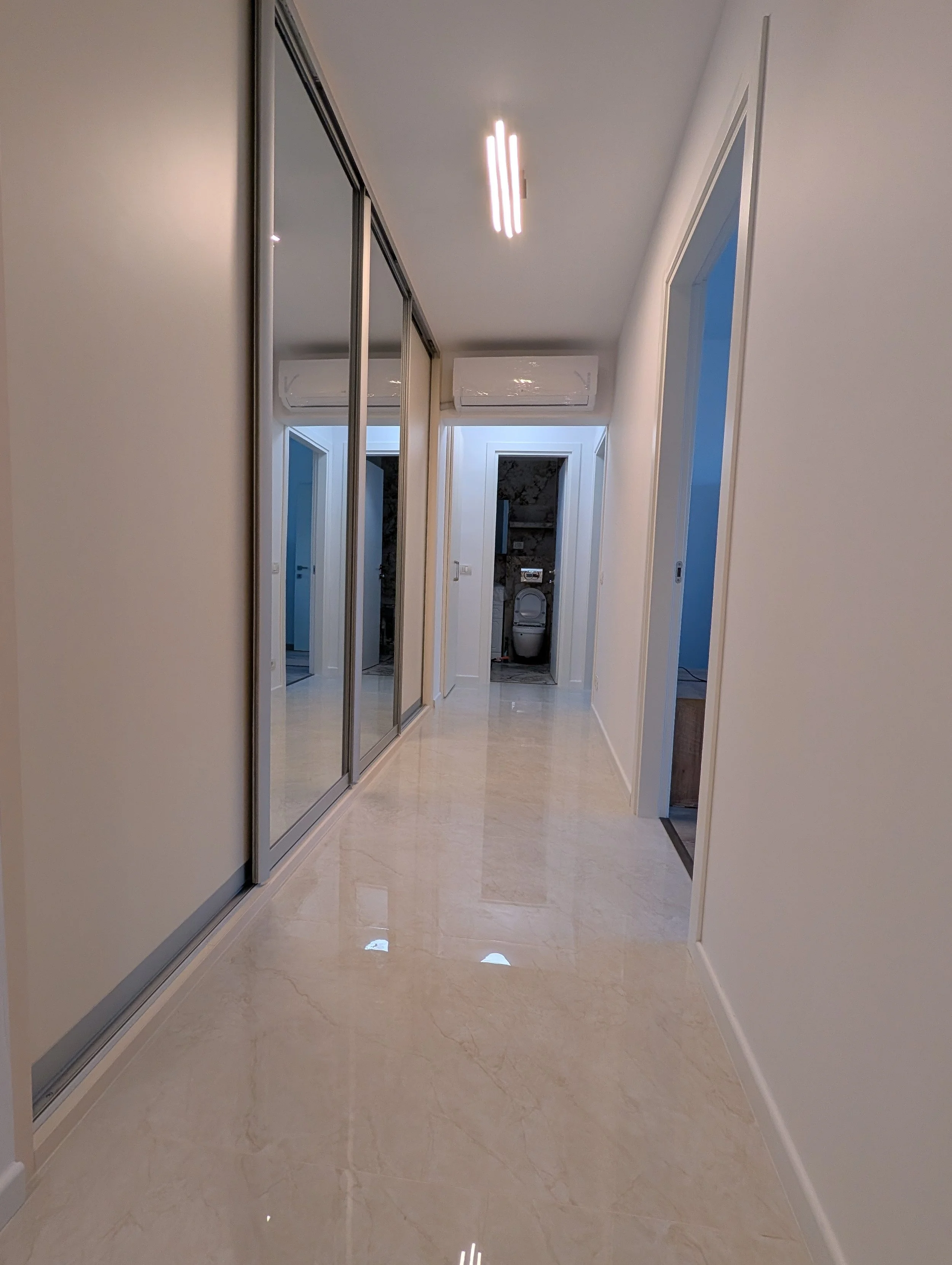 An indoor hallway with polished marble floors, beige walls, mirrored closet doors on the left, and doorways on the right leading to rooms, with a ceiling light and air conditioning unit.