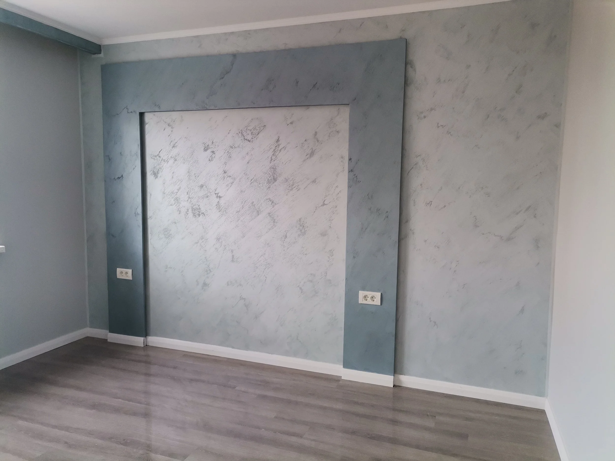 Empty room with gray painted wall, white trim, white ceiling, gray wood flooring, and a double electrical outlet.