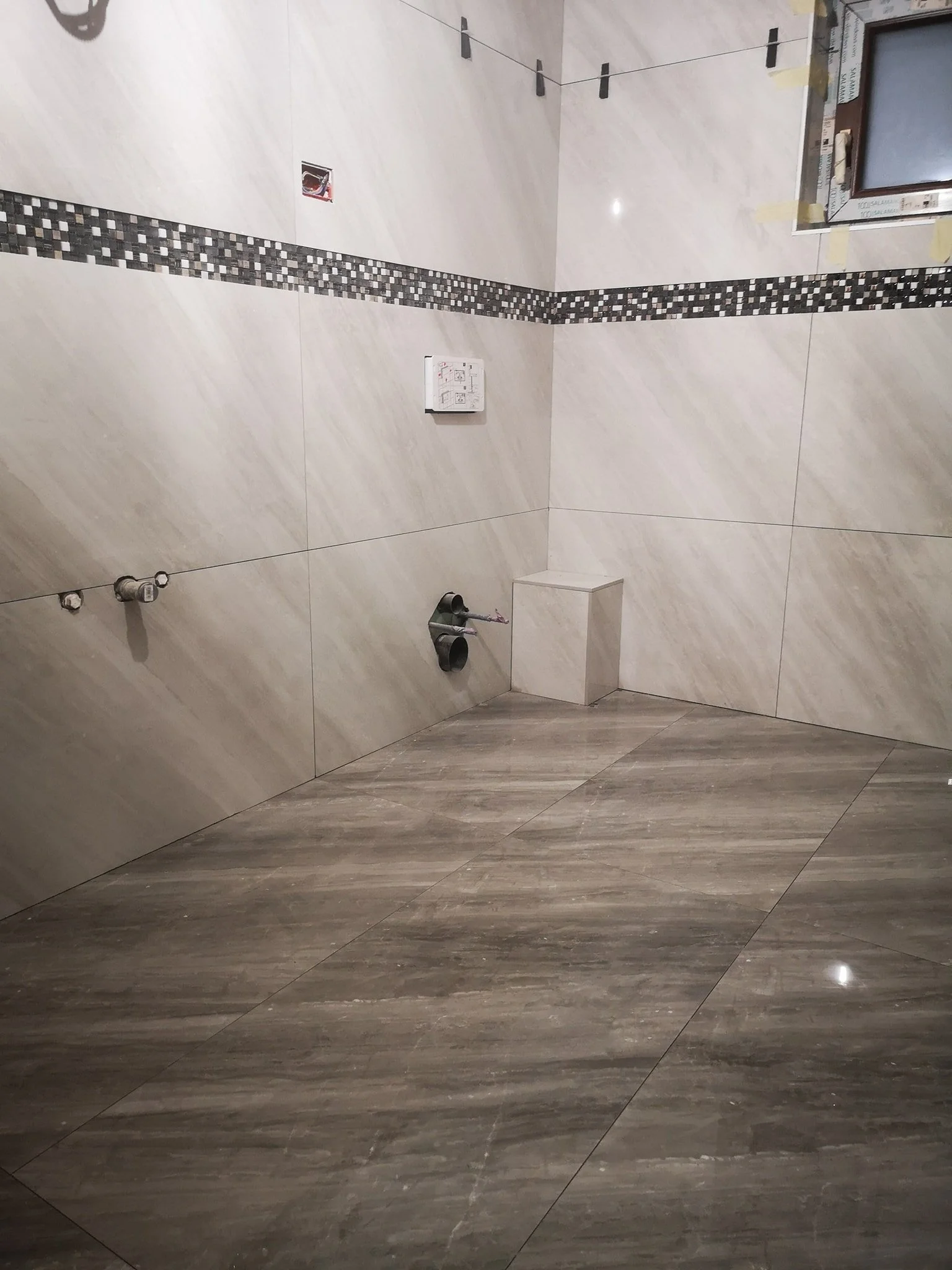 A partially constructed or renovated bathroom with beige and brown tiled walls and floor, an electrical outlet, and plumbing fixtures without a sink or toilet installed yet.