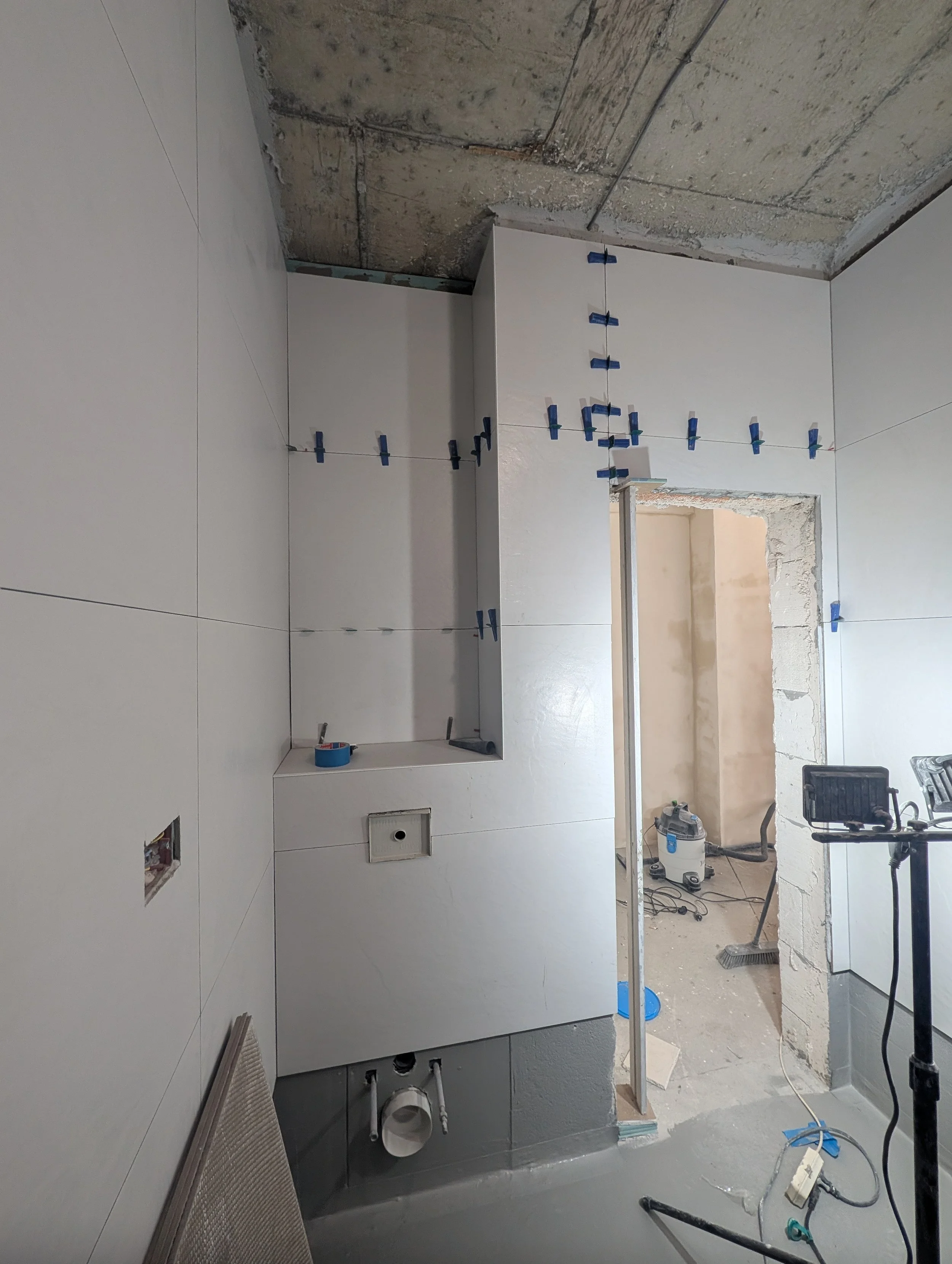 Bathroom under construction with white wall tiles being installed, tools, and equipment visible.